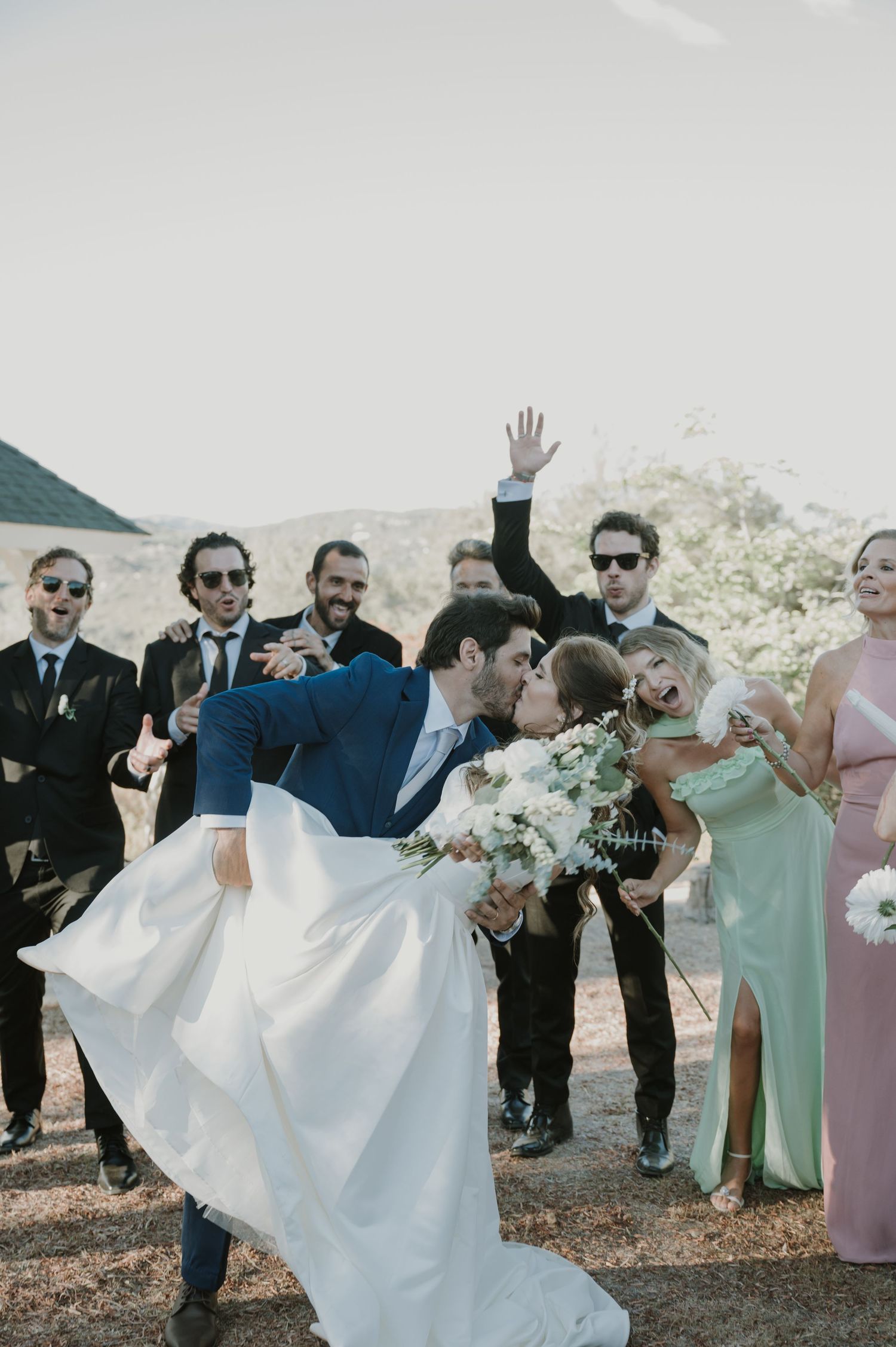Groom dipping bride for a kiss surrounded by cheering wedding party outdoors in scenic setting.