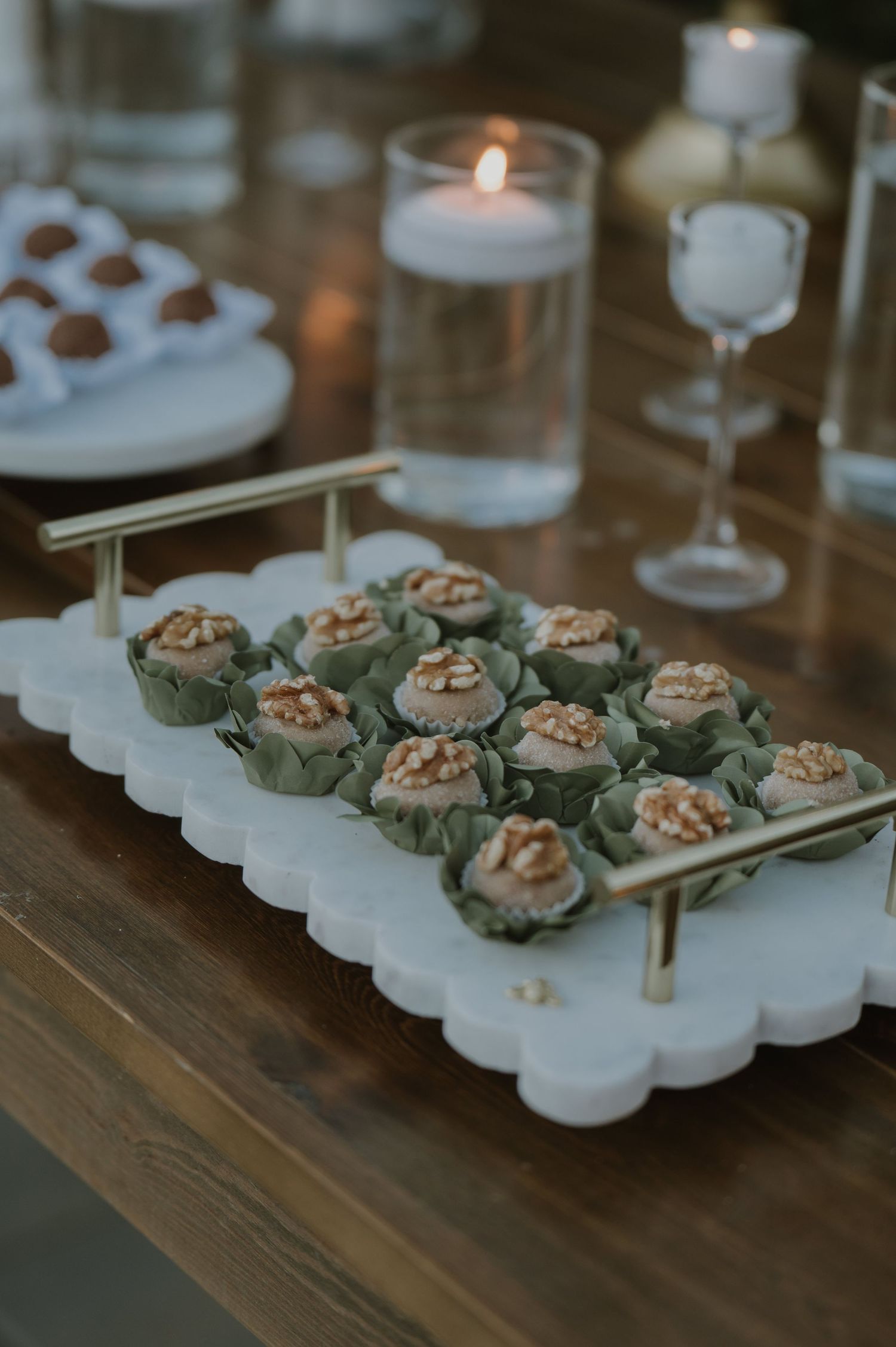Elegant marble tray with gold handles displaying walnut-topped bite-sized desserts in green cups beside candles.