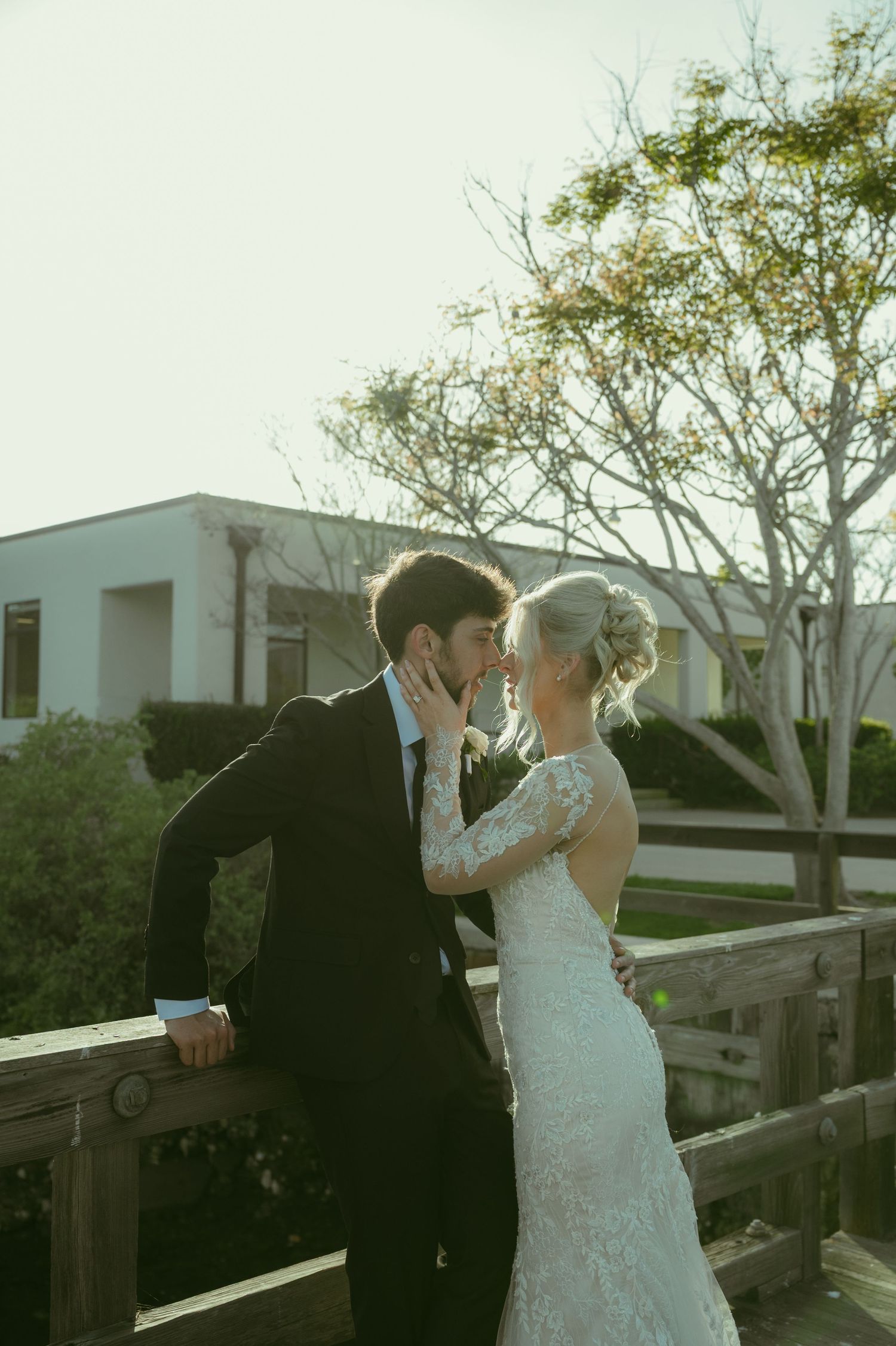 A bride and groom share a romantic kiss on a wooden bridge outdoors, she in lace gown, he in black suit.