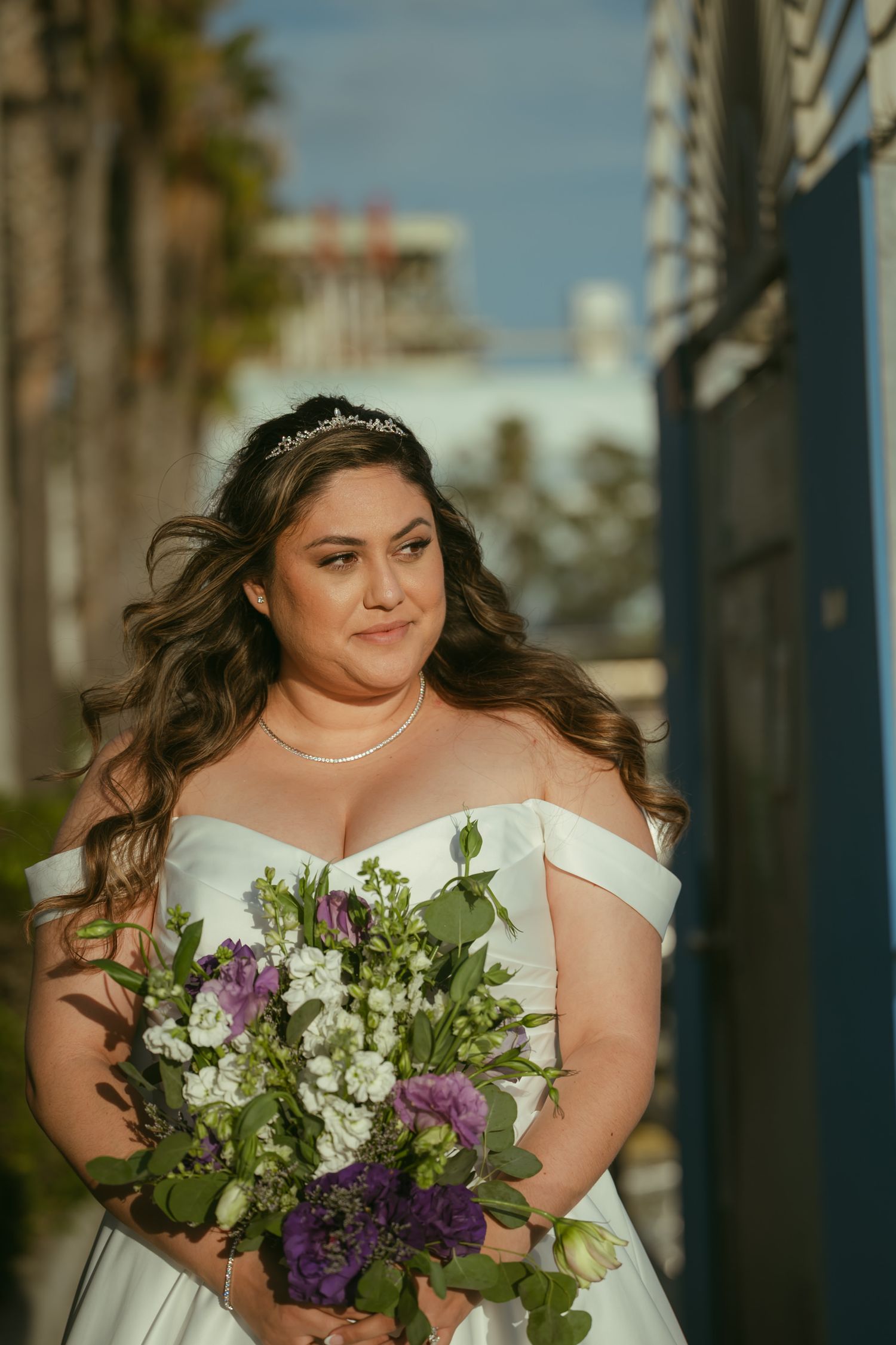 Bride in off-shoulder white gown holding purple and white bouquet, wearing tiara, posing outdoors on sunny day.