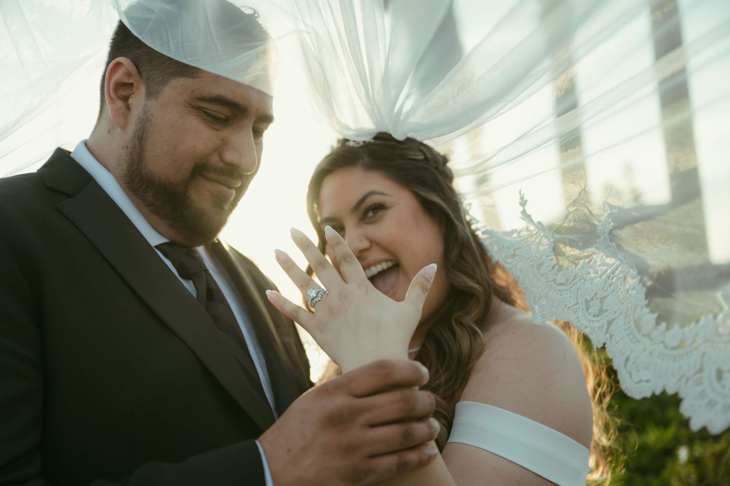 Joyful bride shows off her wedding ring while groom admires it during outdoor ceremony under elegant white draping.
