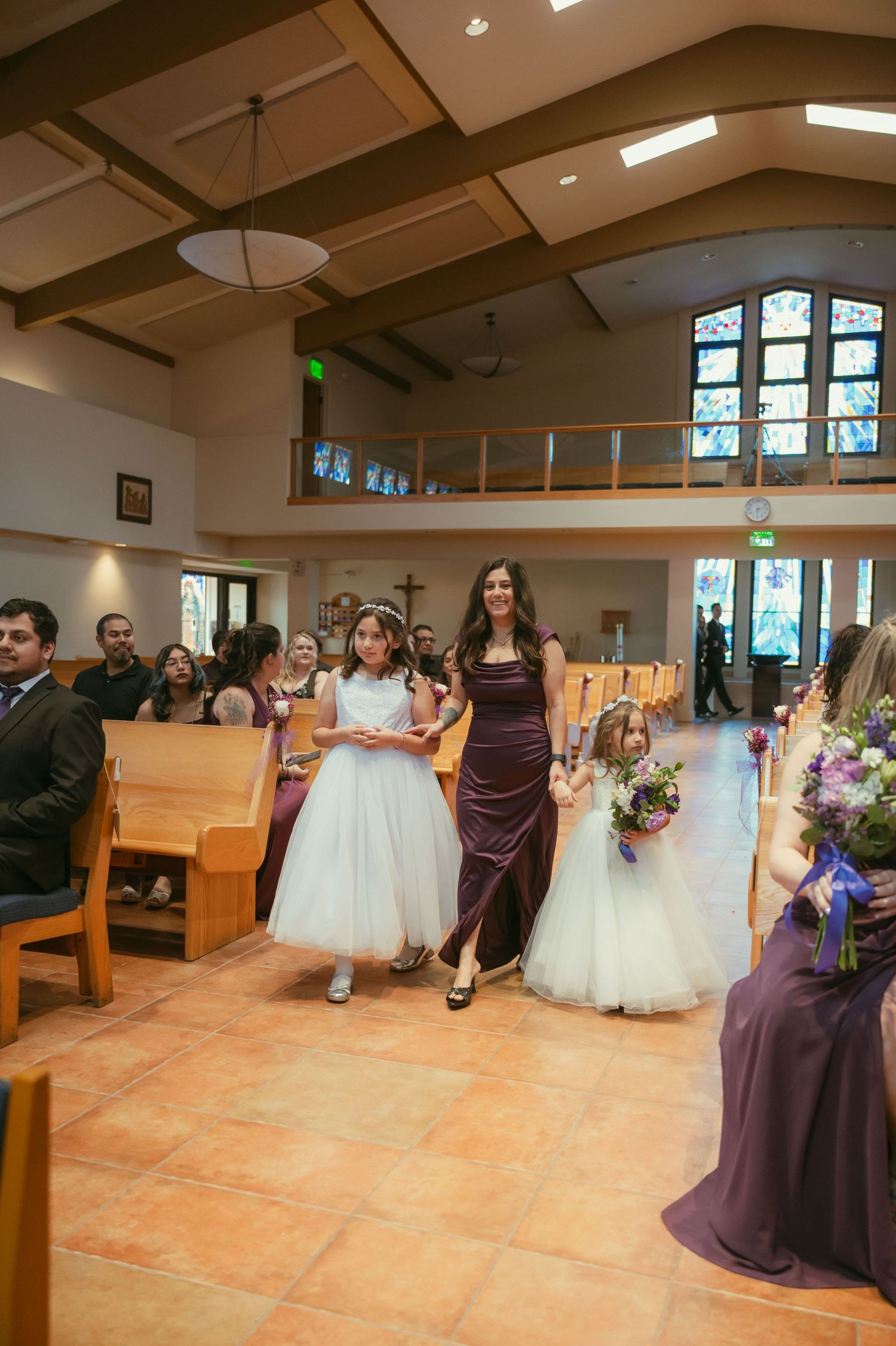 Bridesmaid in purple gown escorts two flower girls in white dresses down church aisle during wedding ceremony.
