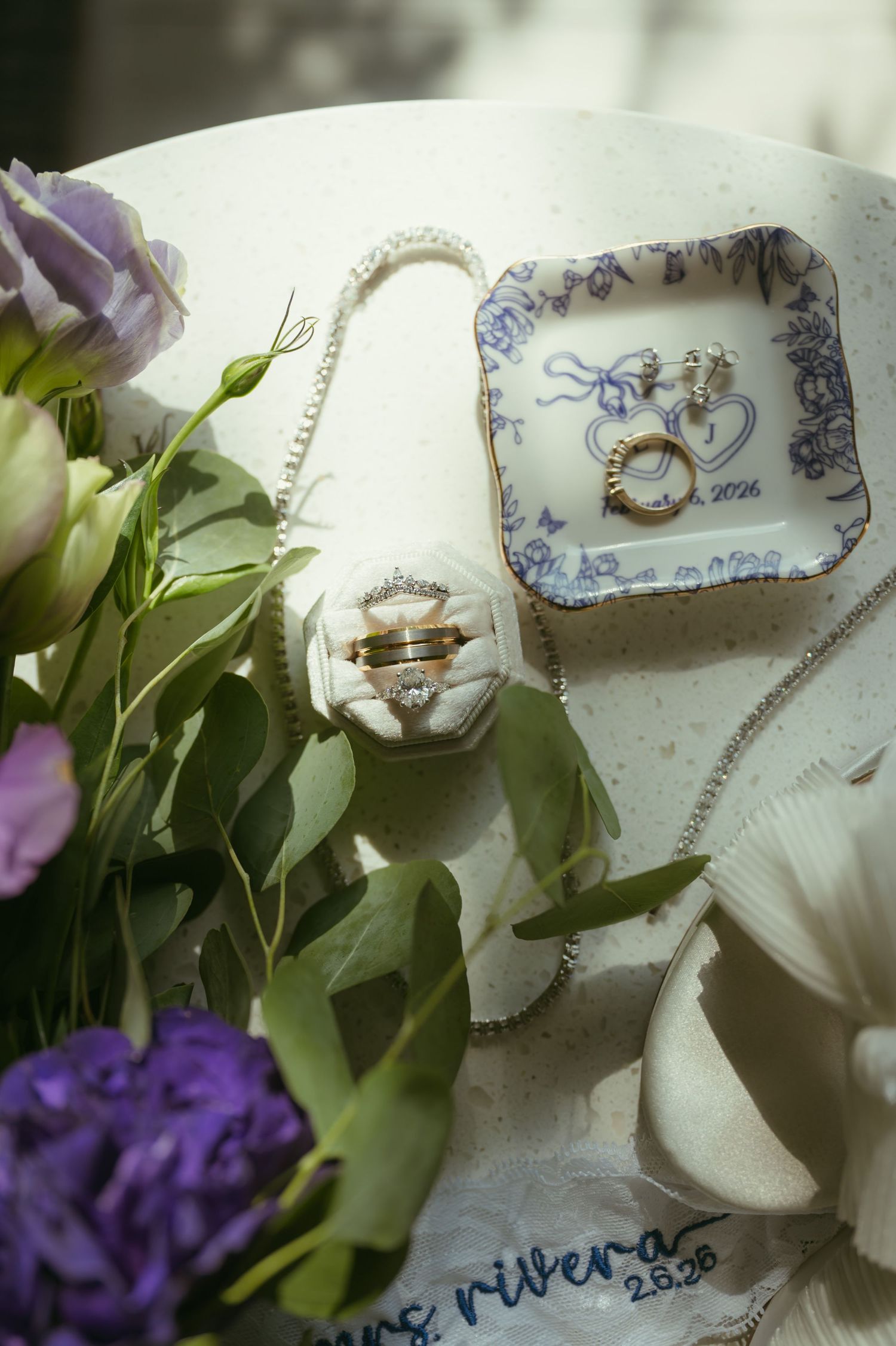 Wedding rings and earrings displayed on elegant ring dish and velvet holder surrounded by purple flowers for Rivera wedding.