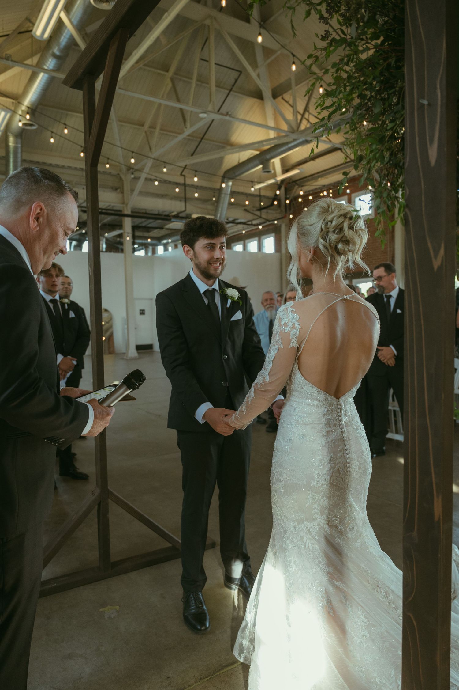 Bride and groom holding hands at indoor wedding ceremony under floral arch with string lights.