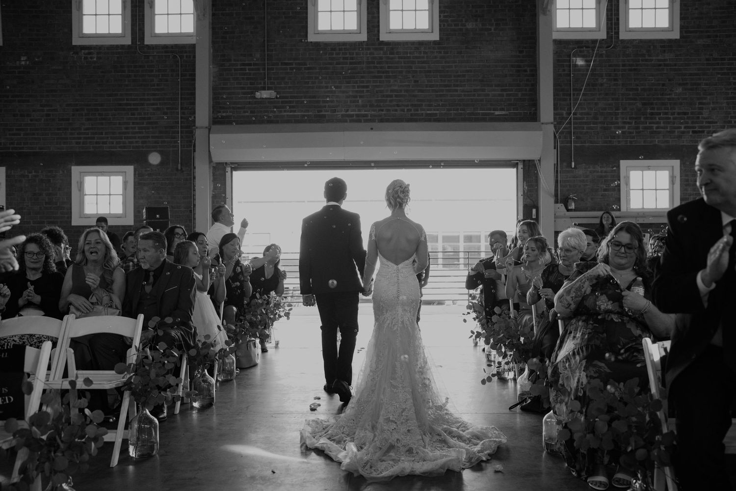 Bride and groom walk hand in hand down aisle after wedding ceremony in industrial venue.
