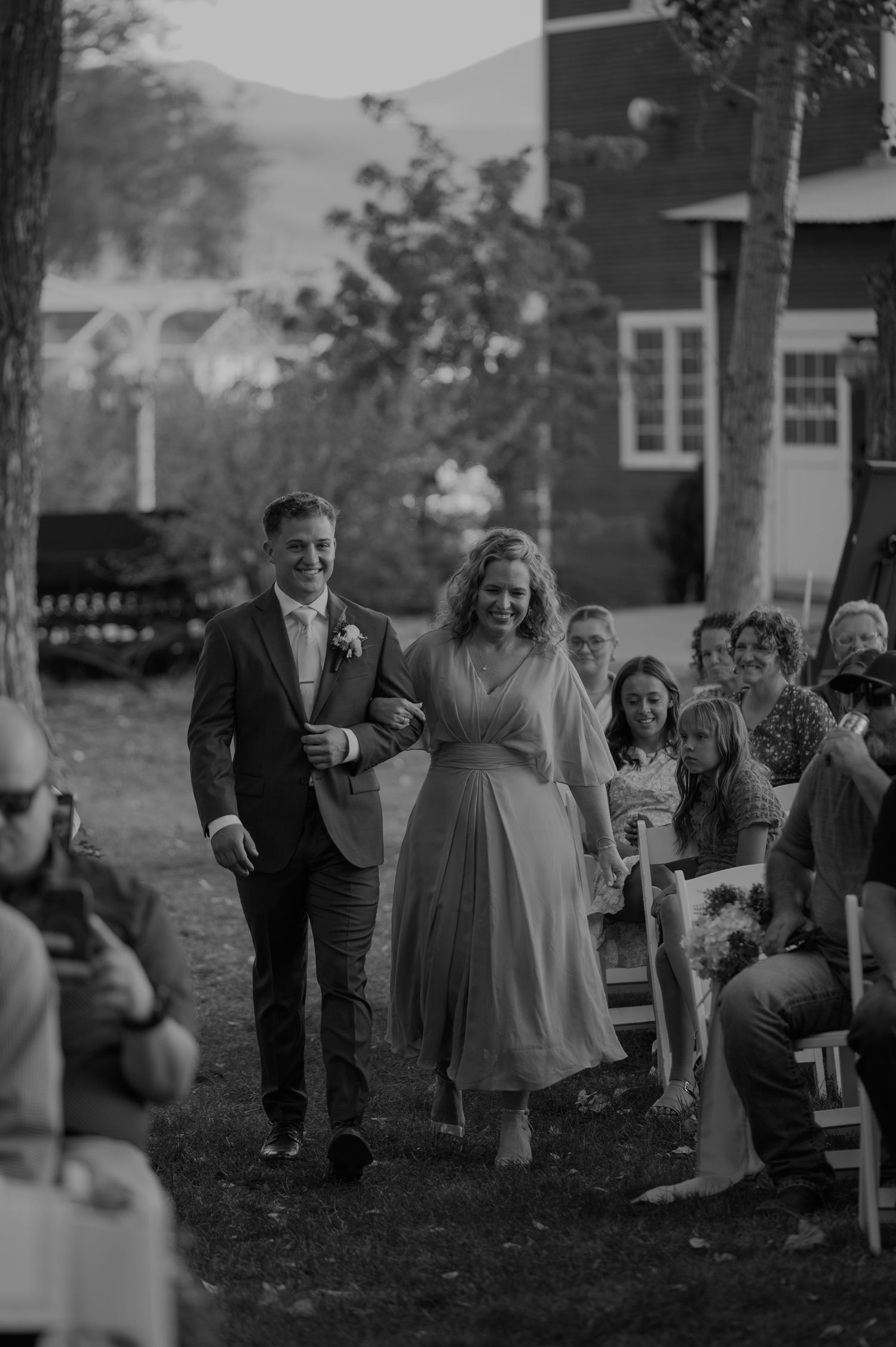 Guests mingling at an outdoor wedding ceremony beneath tall trees.
