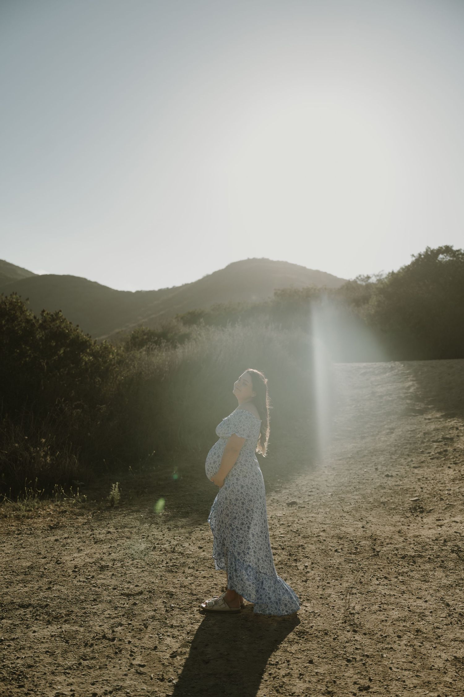 A couple embraces on a hillside during golden hour with mountains in the background.