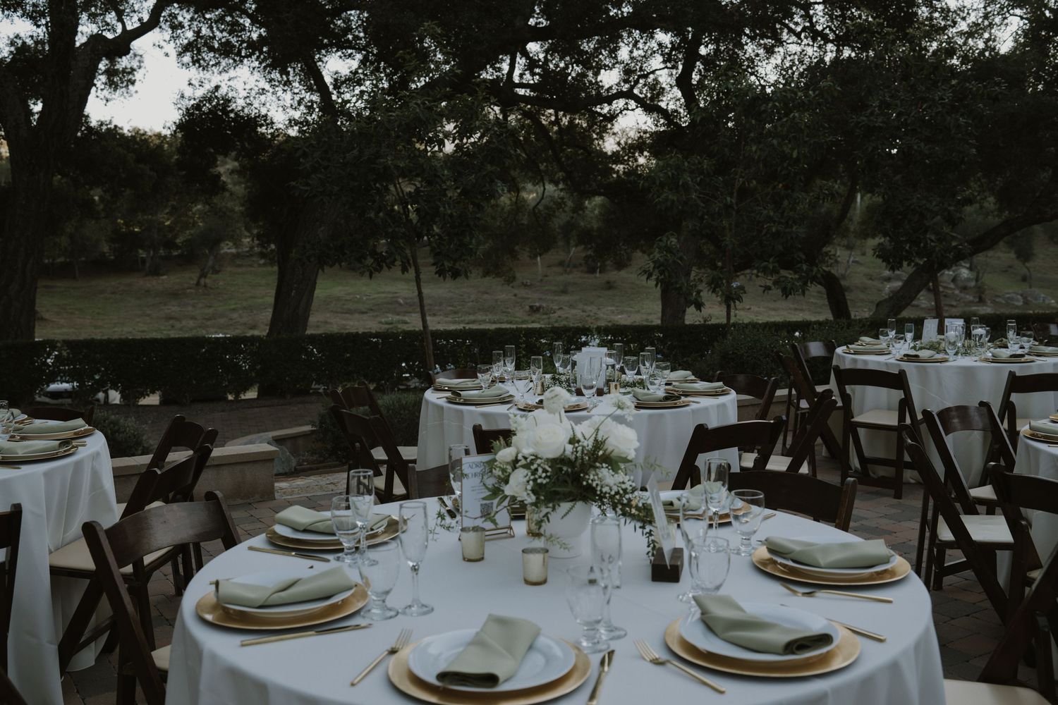 Round dining tables set up outdoors under mature trees for an elegant wedding reception.