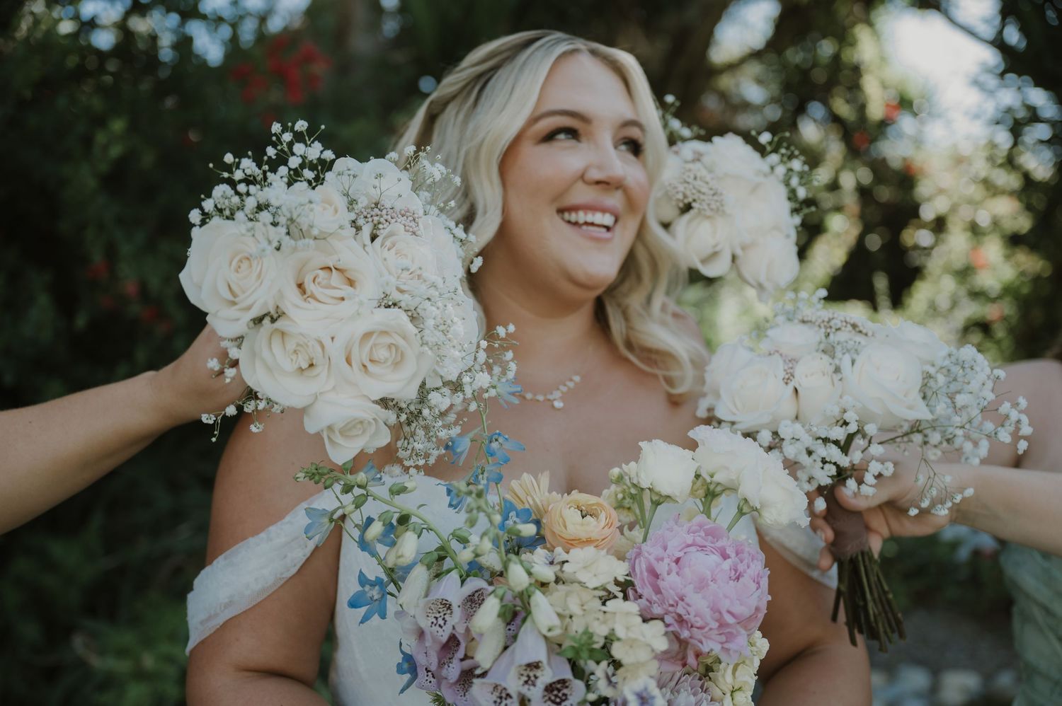 Smiling bride in off-shoulder gown surrounded by white and pink wedding bouquets outdoors.