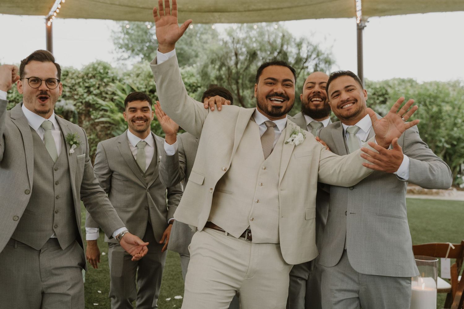 Groom in beige suit celebrating joyfully with groomsmen in gray suits at wedding.