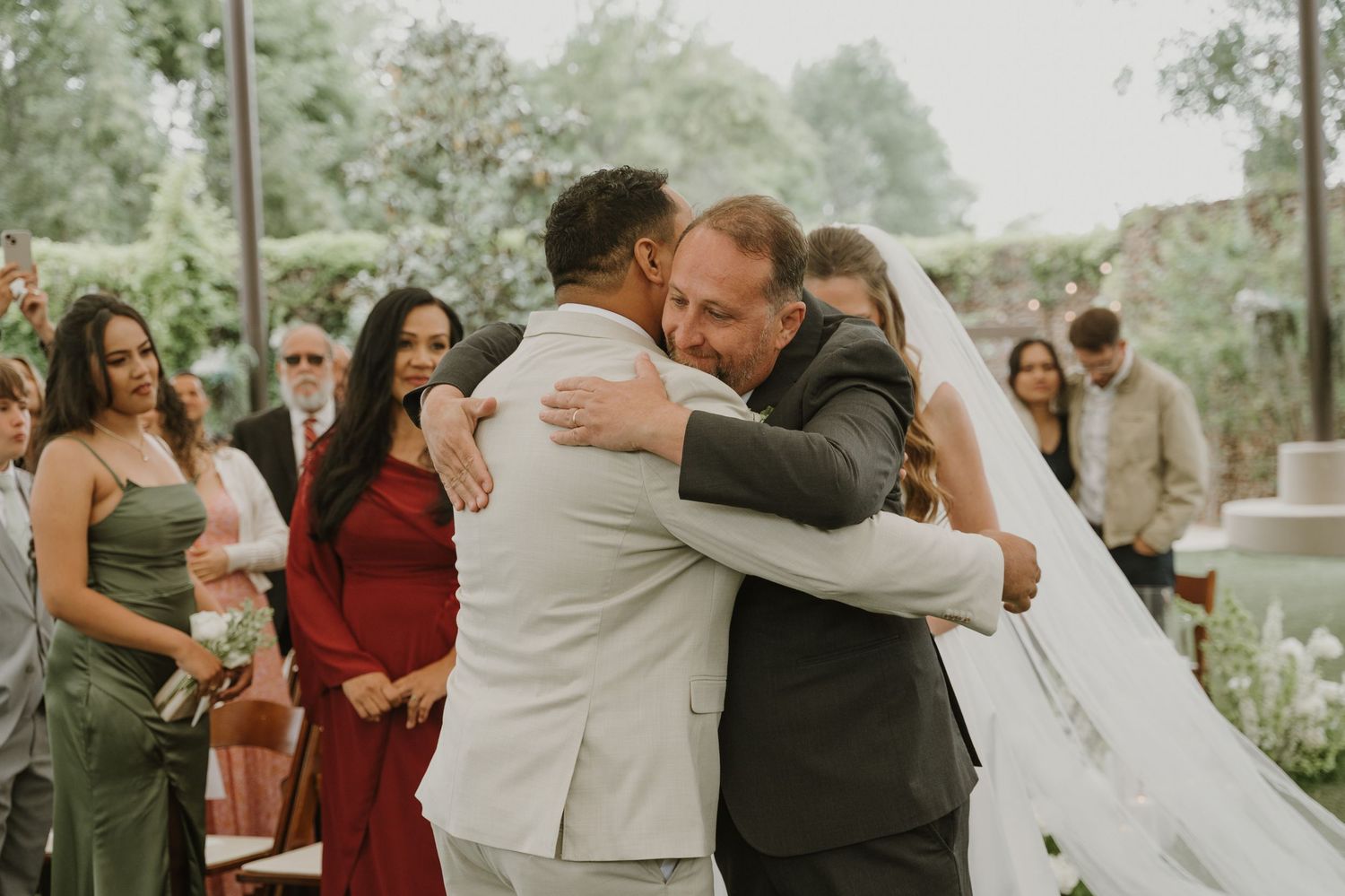 Groom embracing father of the bride during outdoor wedding ceremony while guests watch in background.