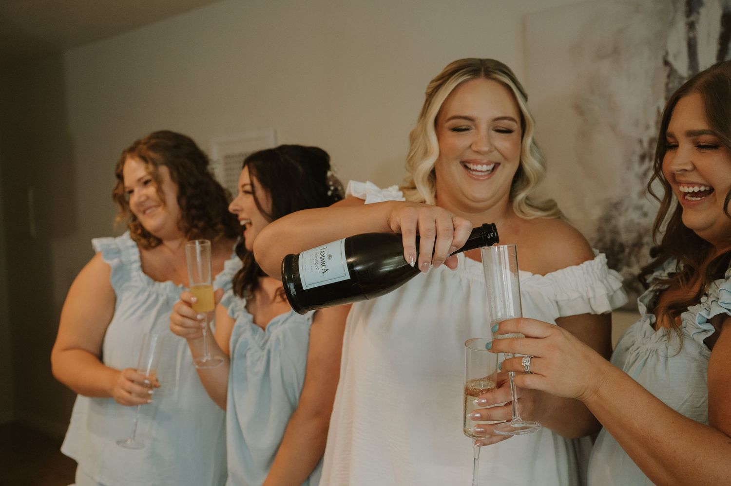 Bride in white off-shoulder dress pours champagne while bridesmaids in light blue dresses celebrate together.