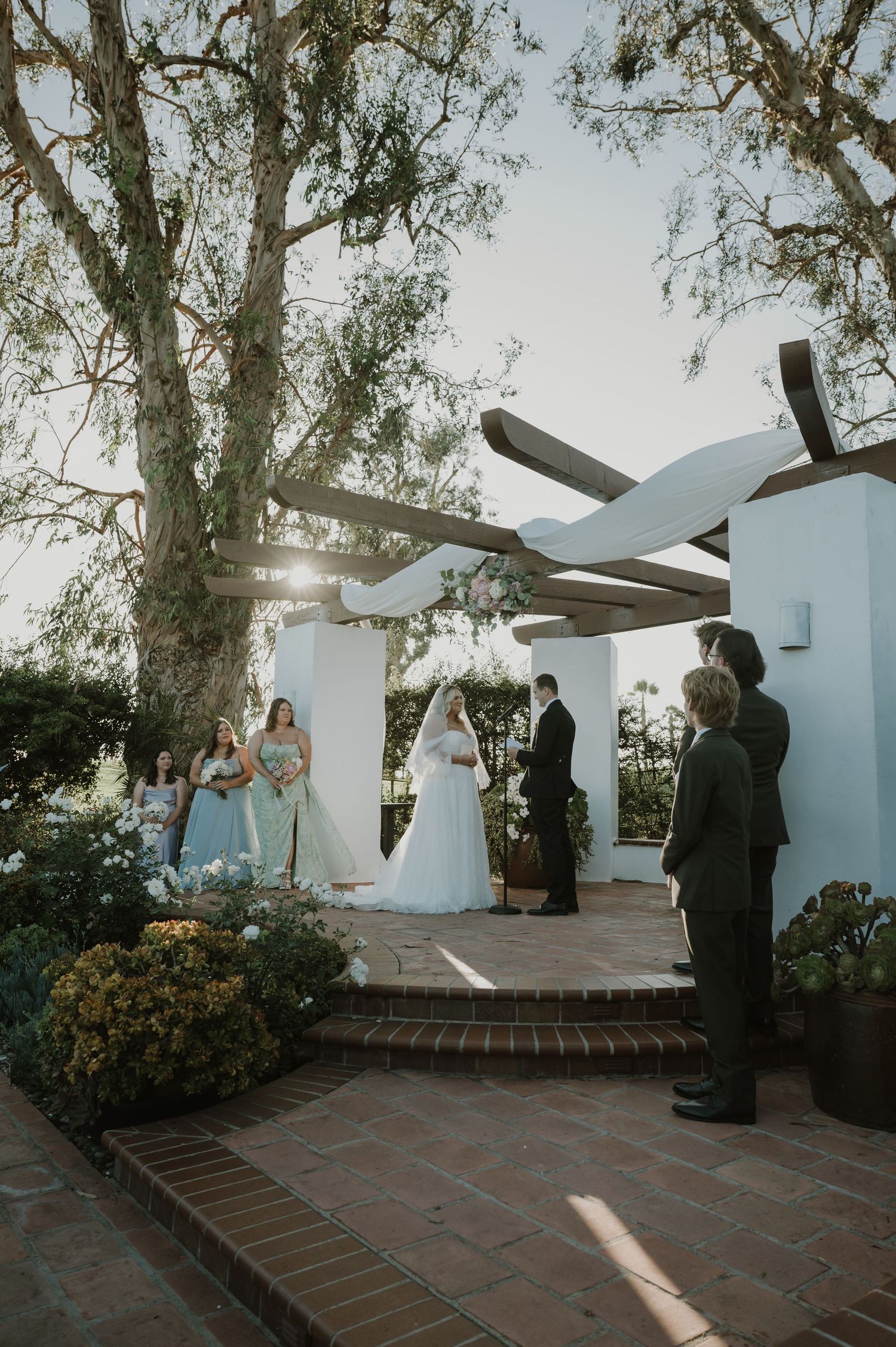 Outdoor wedding ceremony under a rustic pergola with bride, groom, officiant, and bridesmaids in blue dresses amid lush gardens.