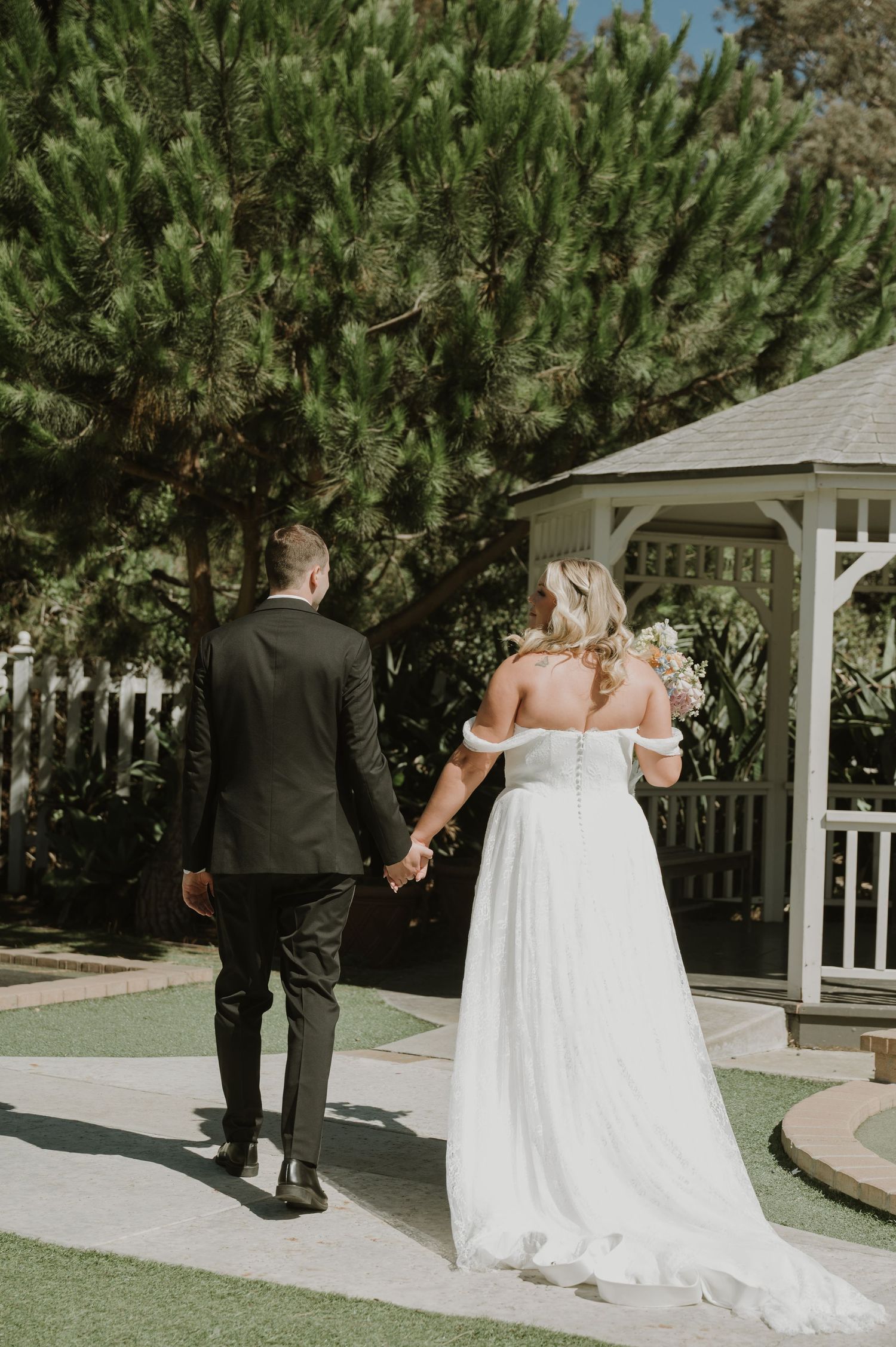 A bride and groom walk hand in hand toward a gazebo surrounded by lush green trees on their wedding day.