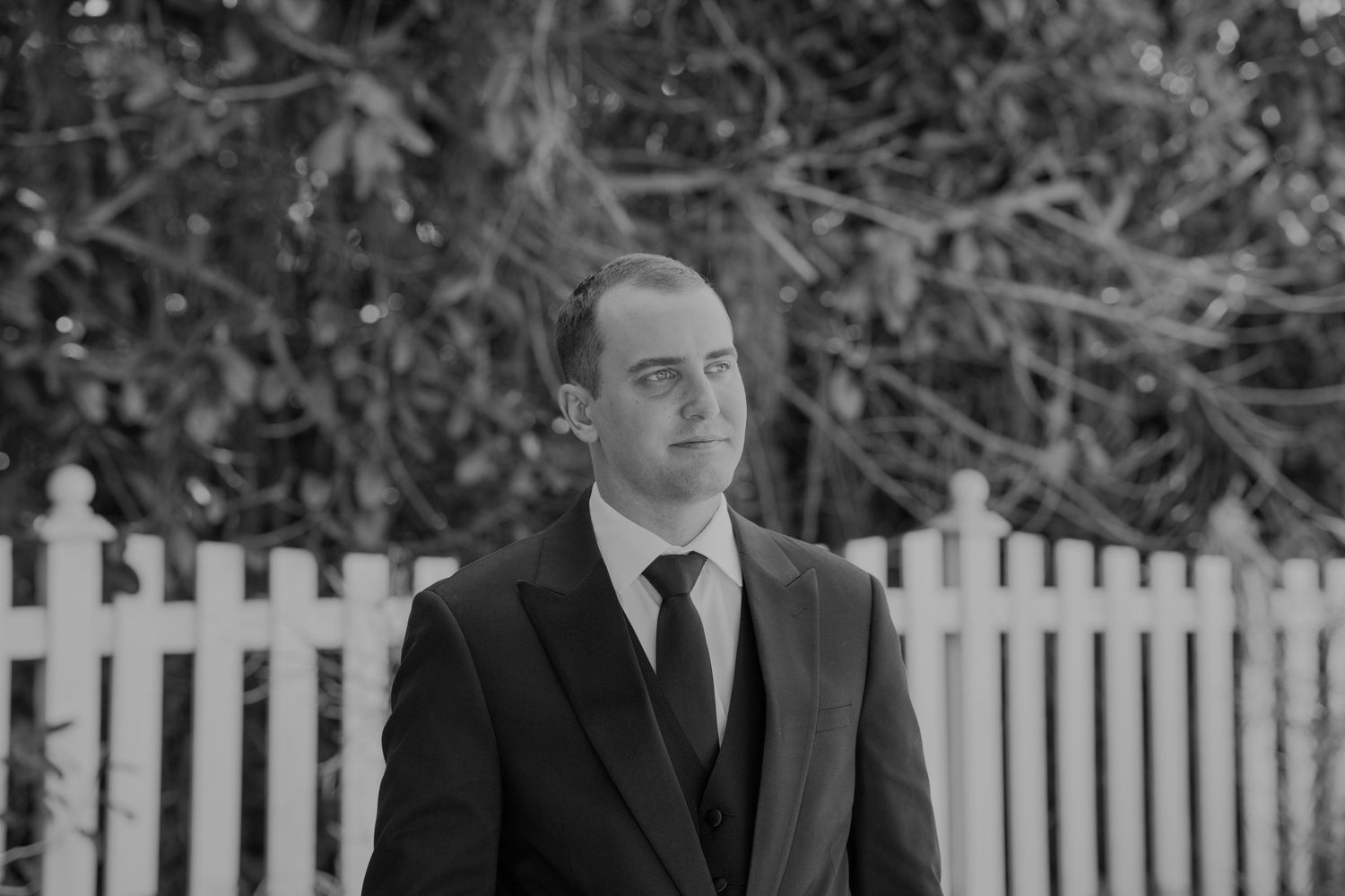 Man in dark suit and tie standing outdoors near white picket fence, black and white portrait photo.