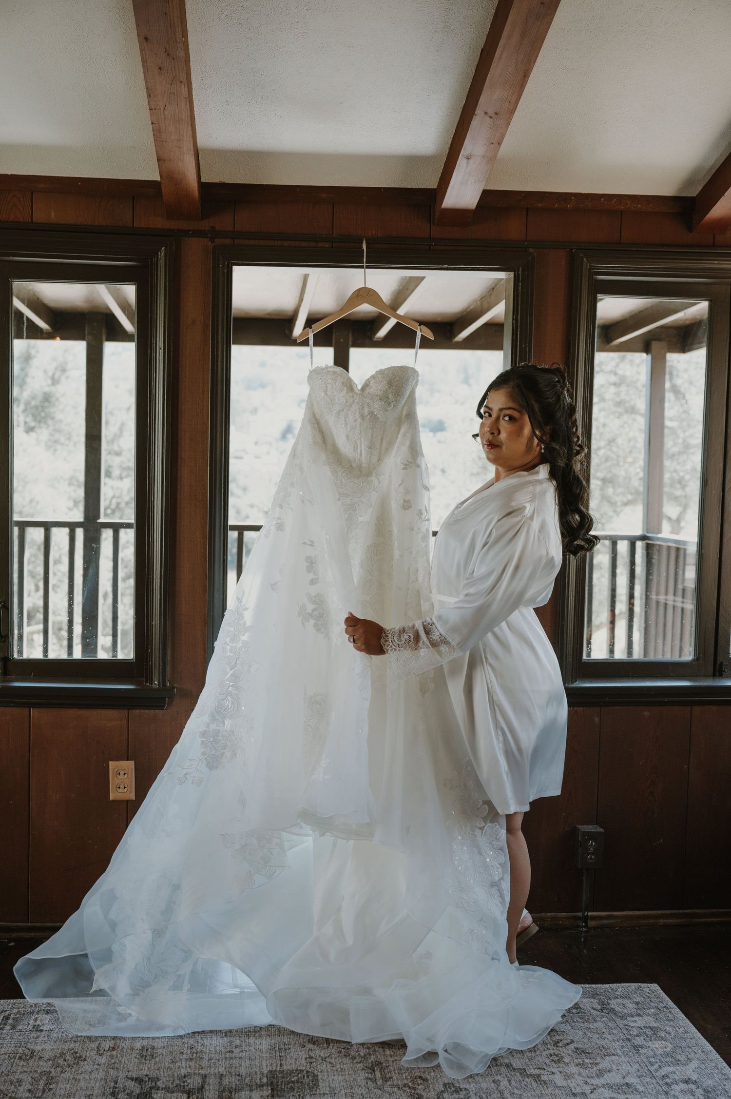 Bride in white robe standing before her elegant strapless wedding gown hanging in a rustic wood-paneled room.