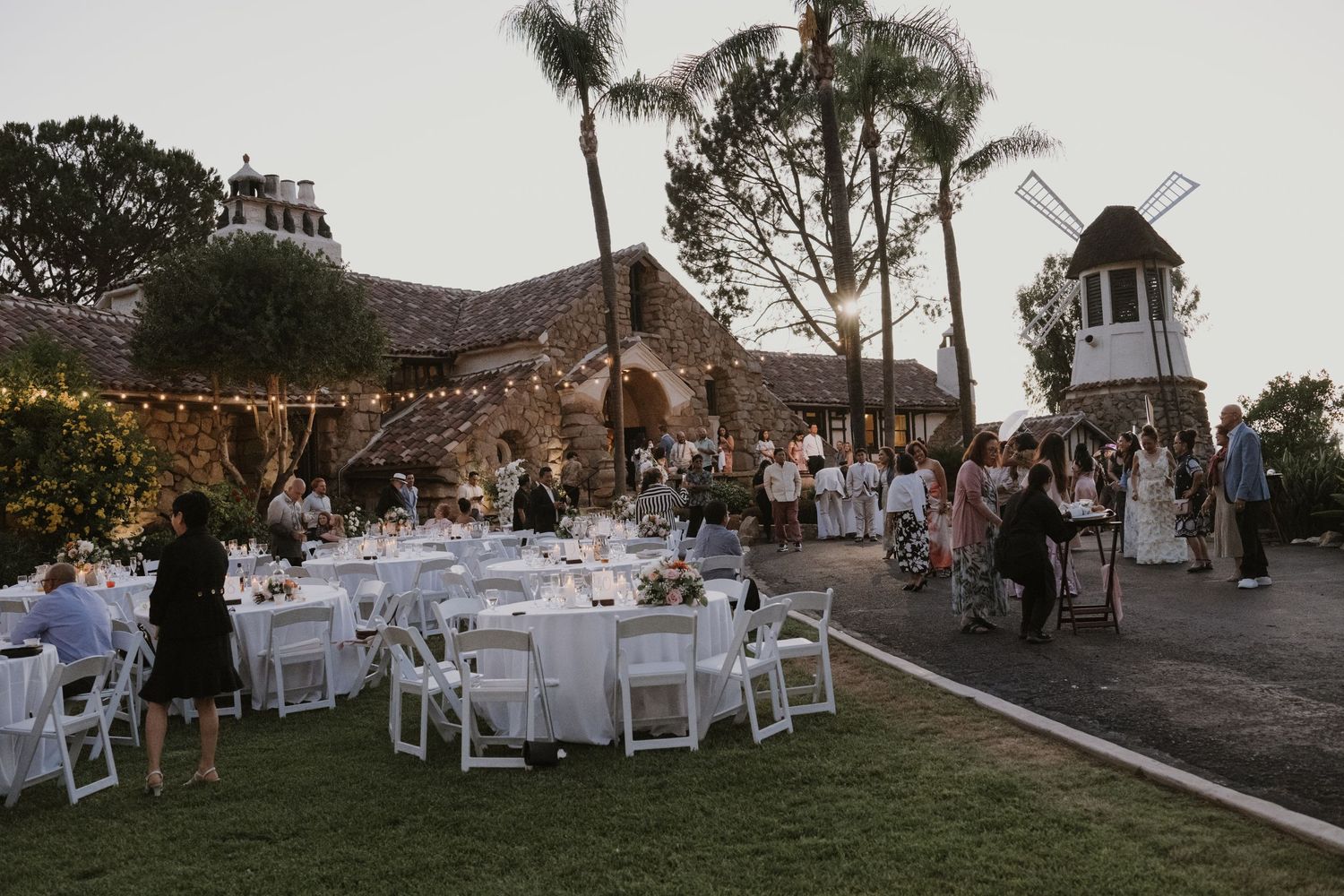 Elegant outdoor wedding reception at dusk featuring white round tables, string lights, and a scenic windmill backdrop.