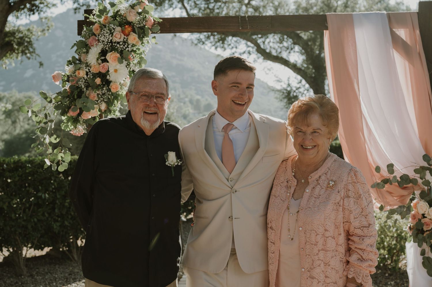 Groom in beige suit poses with elderly couple at floral wedding altar with mountain backdrop.