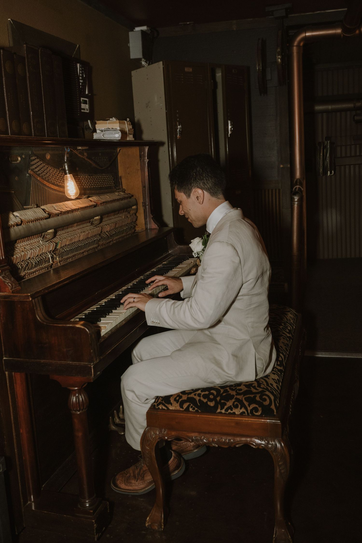 Groom in white wedding suit plays antique upright piano in warmly lit room.