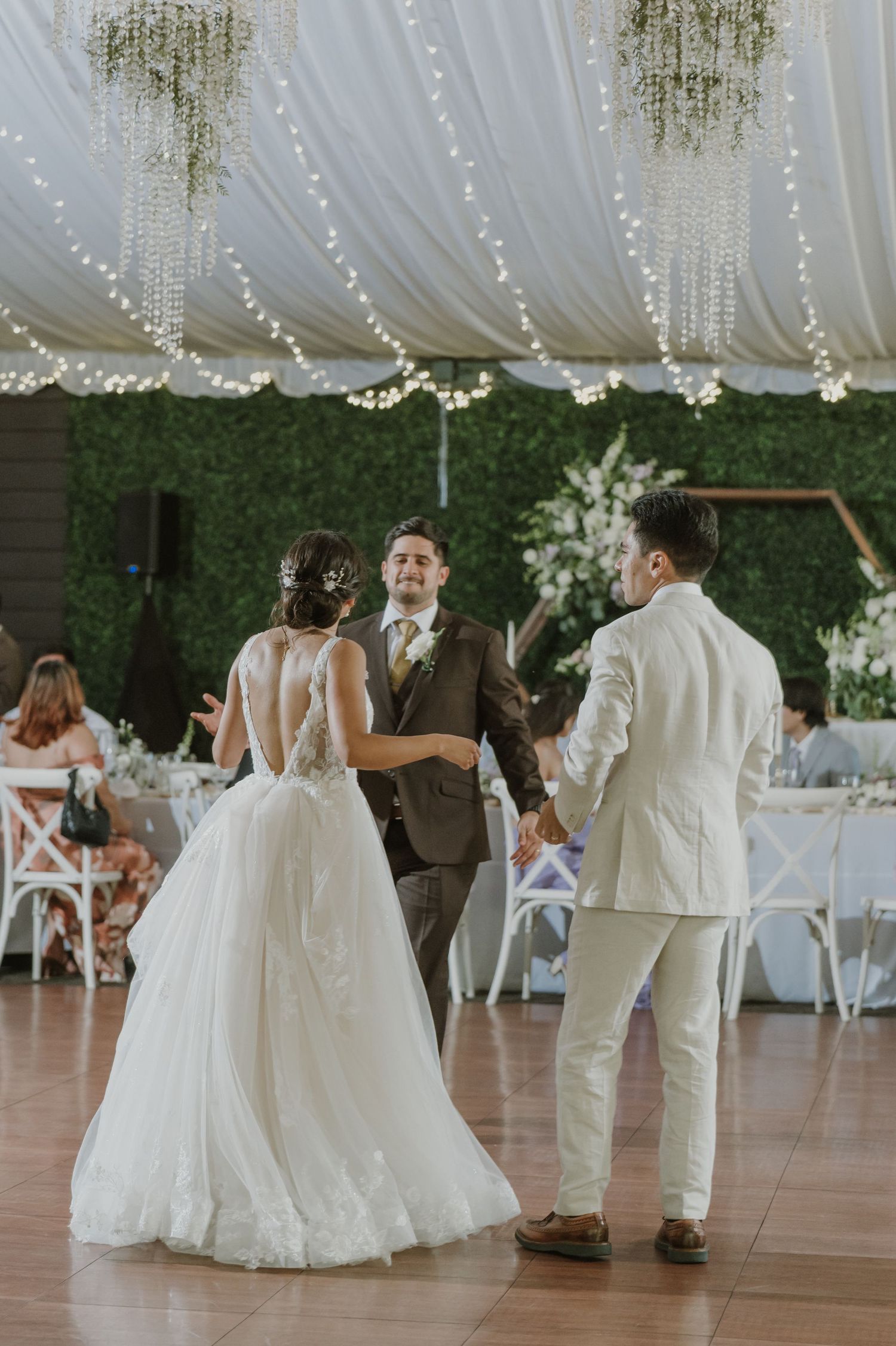 Bride in a backless lace gown dances with two men under fairy lights in a greenery-adorned reception tent.