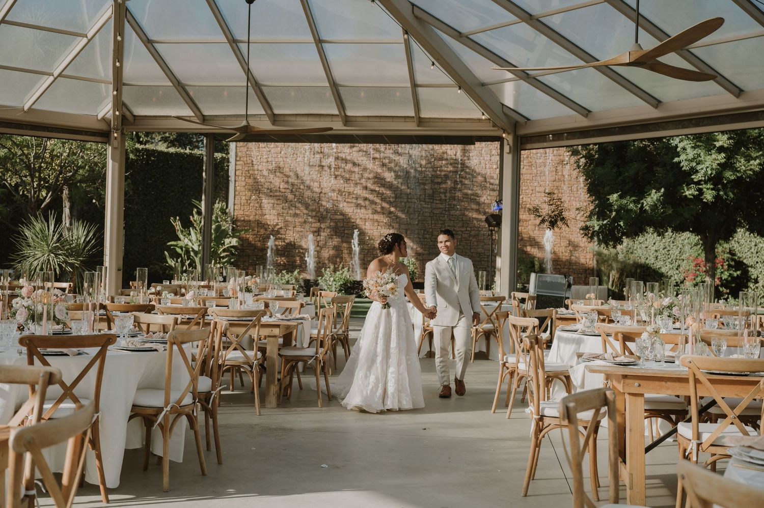 Bride and groom stand together in an elegant glass greenhouse wedding venue with wooden chairs and white table settings.