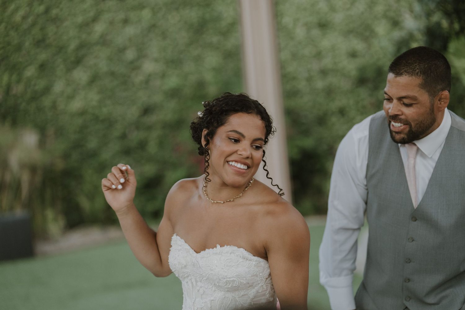 Joyful bride laughs and dances with smiling groom in gray vest at outdoor wedding celebration.