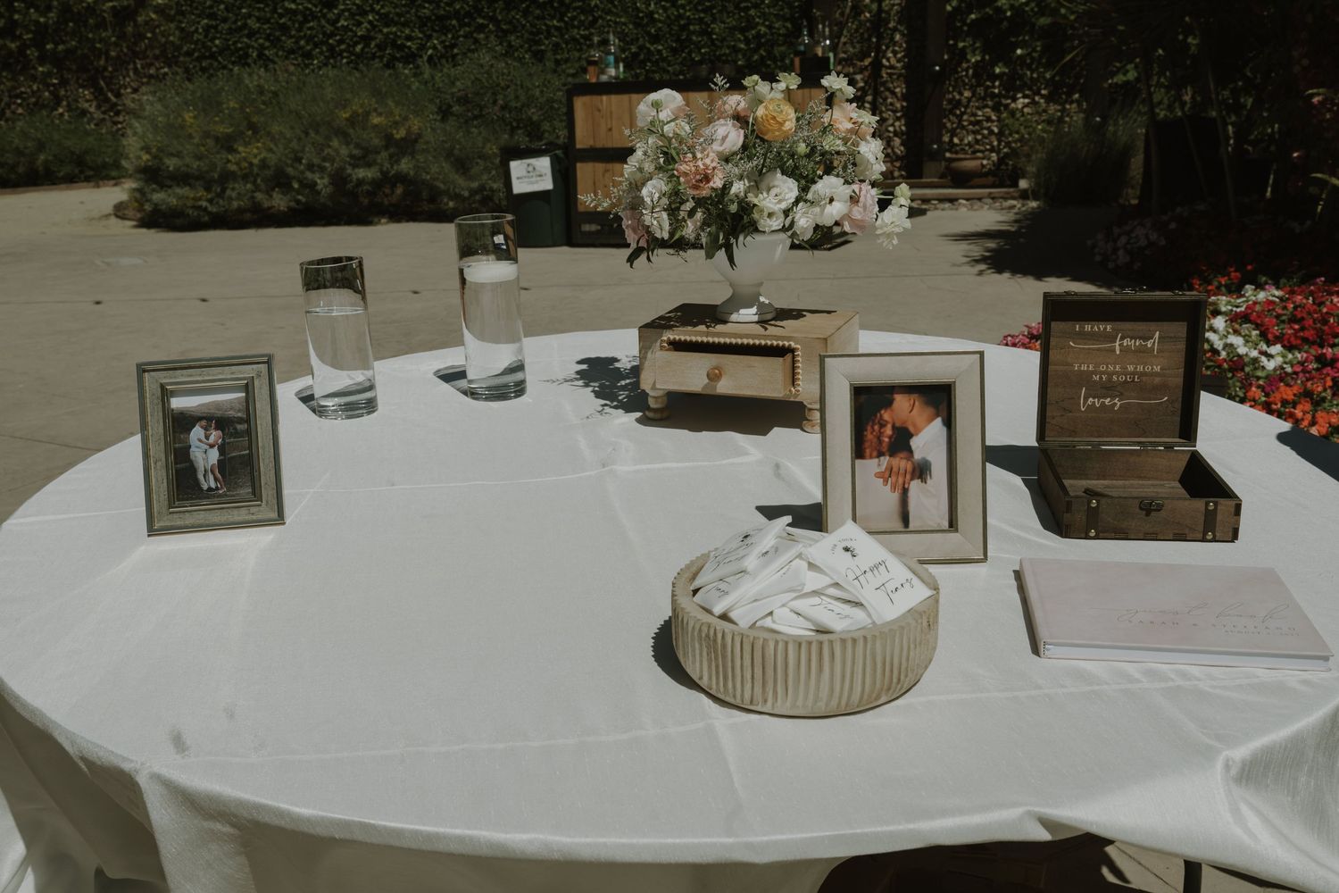 Outdoor wedding welcome table with framed photos, guest book, and floral centerpiece on white linen.