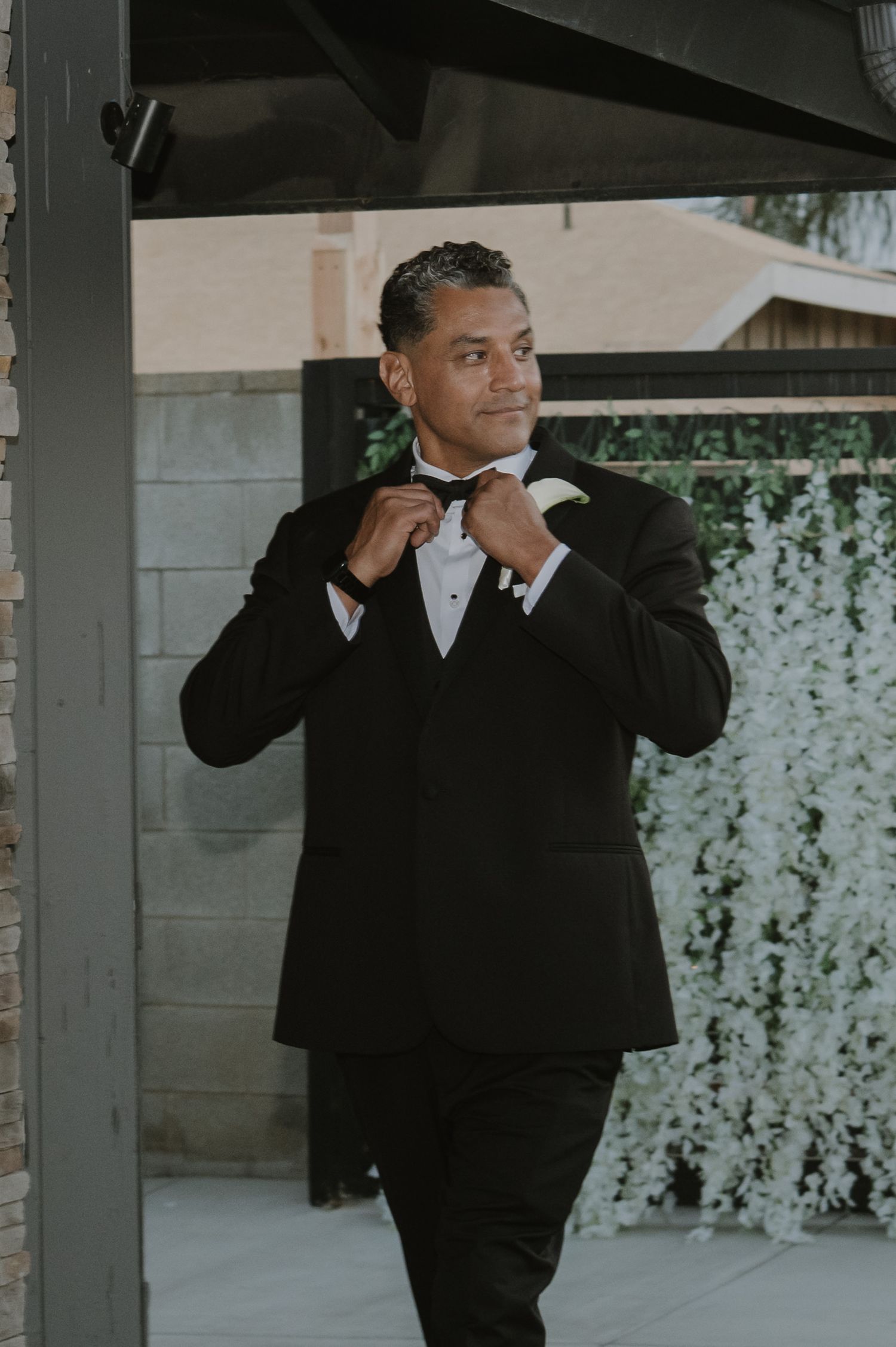 A groom in a black tuxedo adjusts his bow tie outdoors near a white floral wall backdrop.