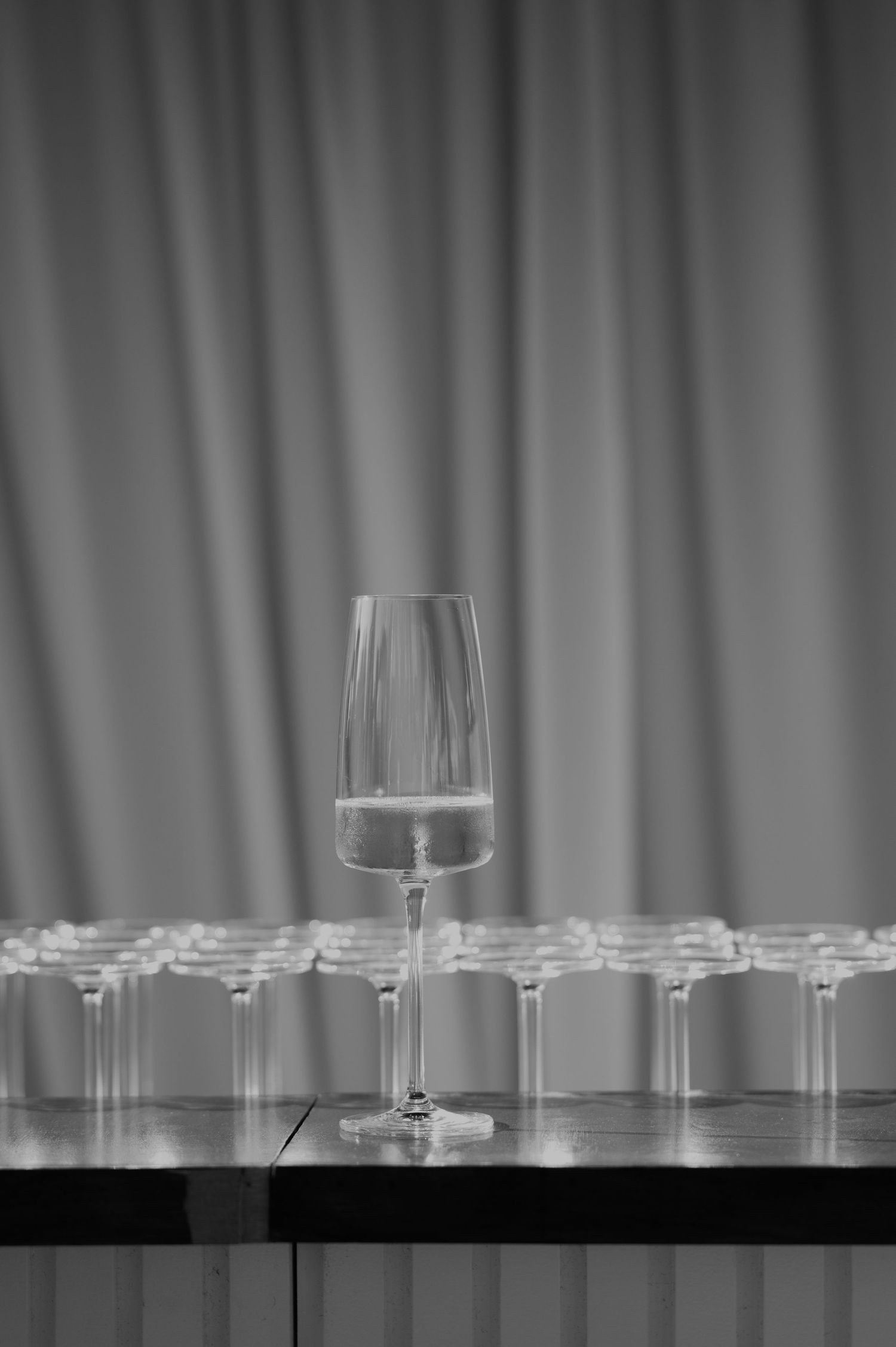 Row of empty champagne flutes on table with single glass in focus against draped curtain backdrop.
