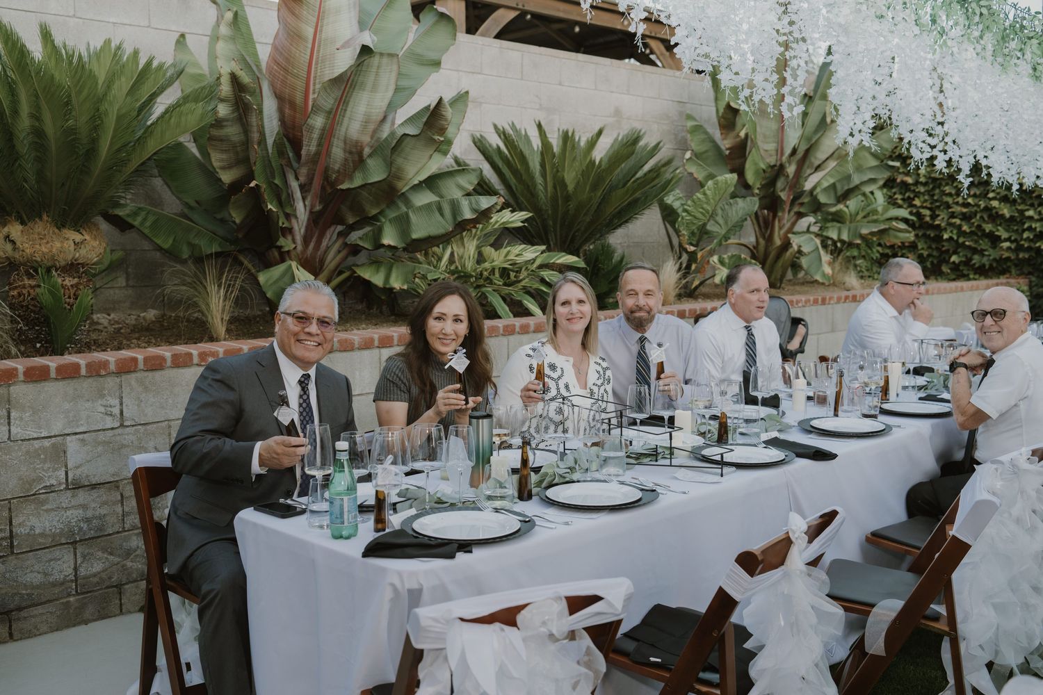 Guests smiling and seated at an elegant outdoor wedding reception table with white linens and tropical greenery backdrop.