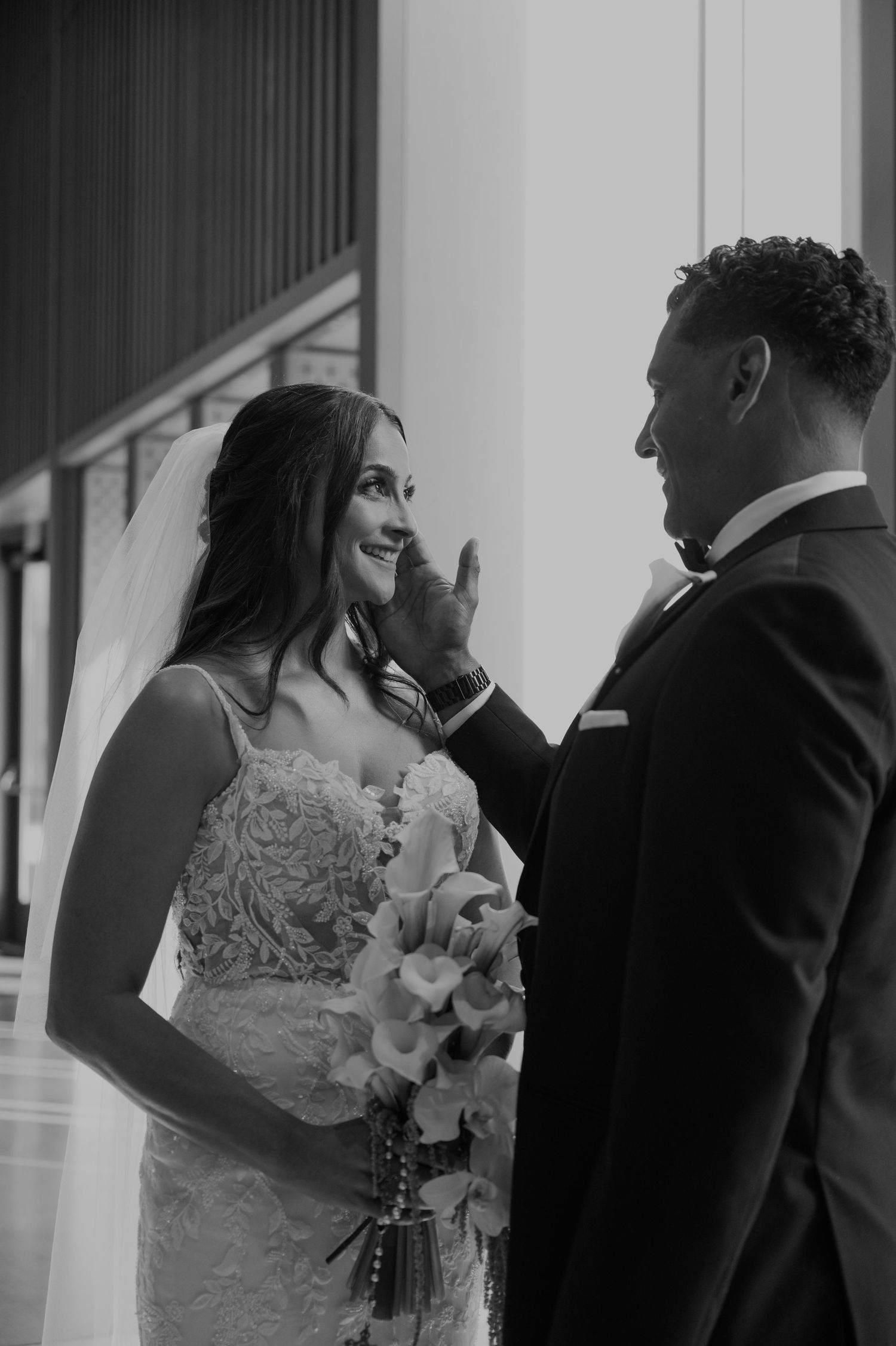 Black and white photo of a groom tenderly touching his bride's face as she smiles holding a floral bouquet on their wedding day.