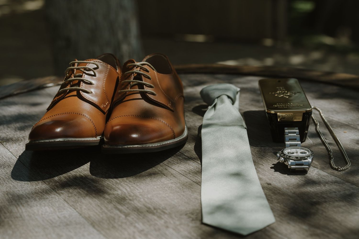 Brown leather oxford shoes beside a sage green tie, gold belt buckle, and wristwatch on a wooden surface.