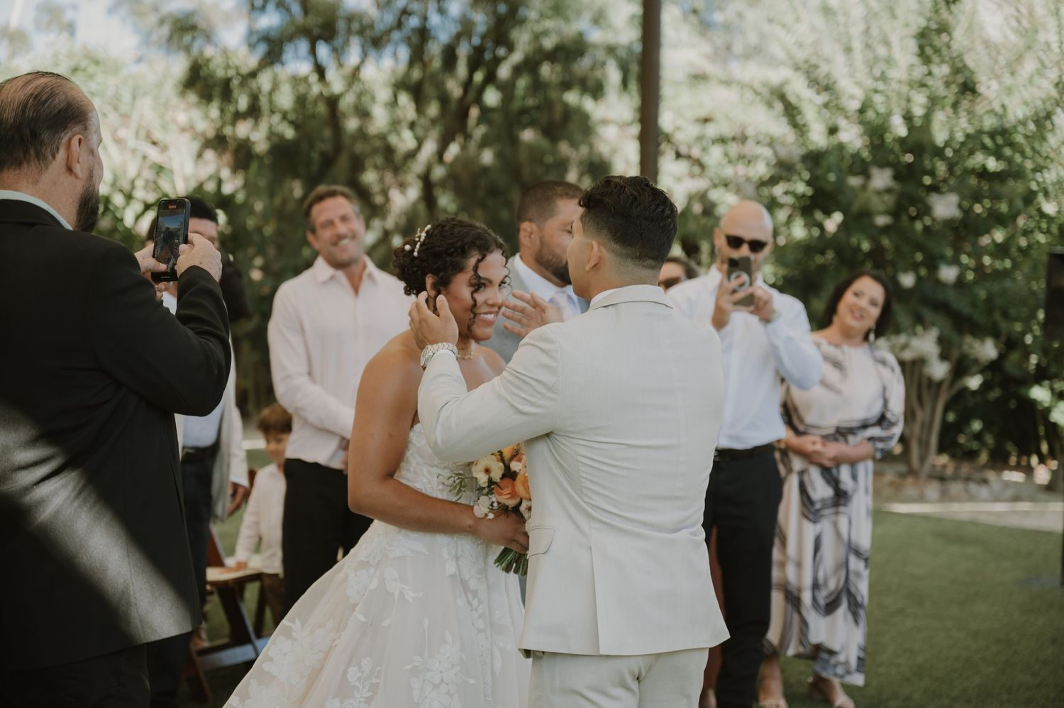 Bride and groom share first kiss surrounded by guests at outdoor garden wedding ceremony.