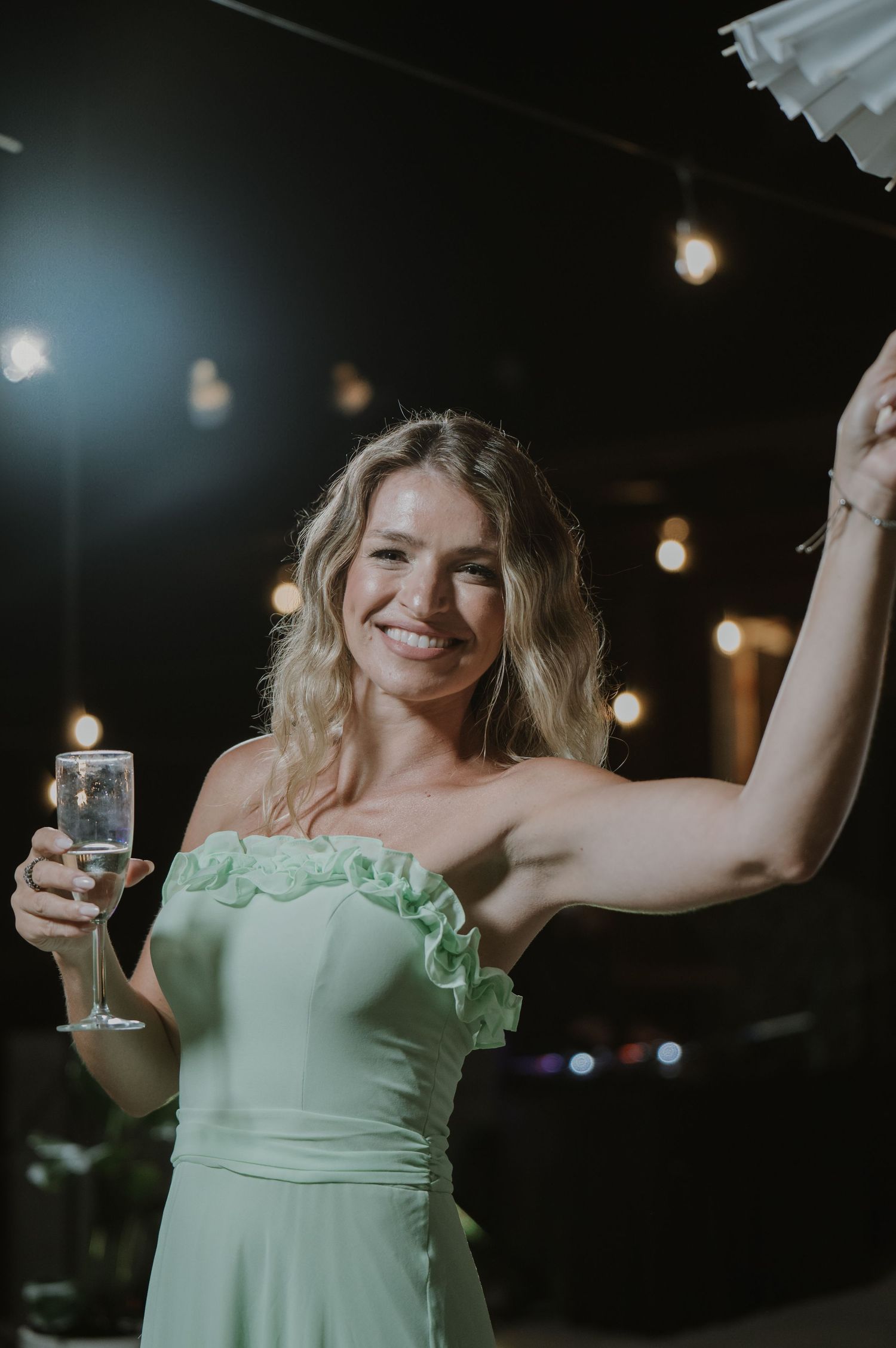 Smiling bridesmaid in mint green strapless dress holding champagne glass at evening wedding reception.