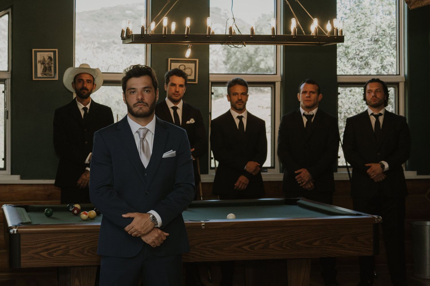 Groom in navy suit poses with groomsmen near a pool table in a rustic wedding venue.