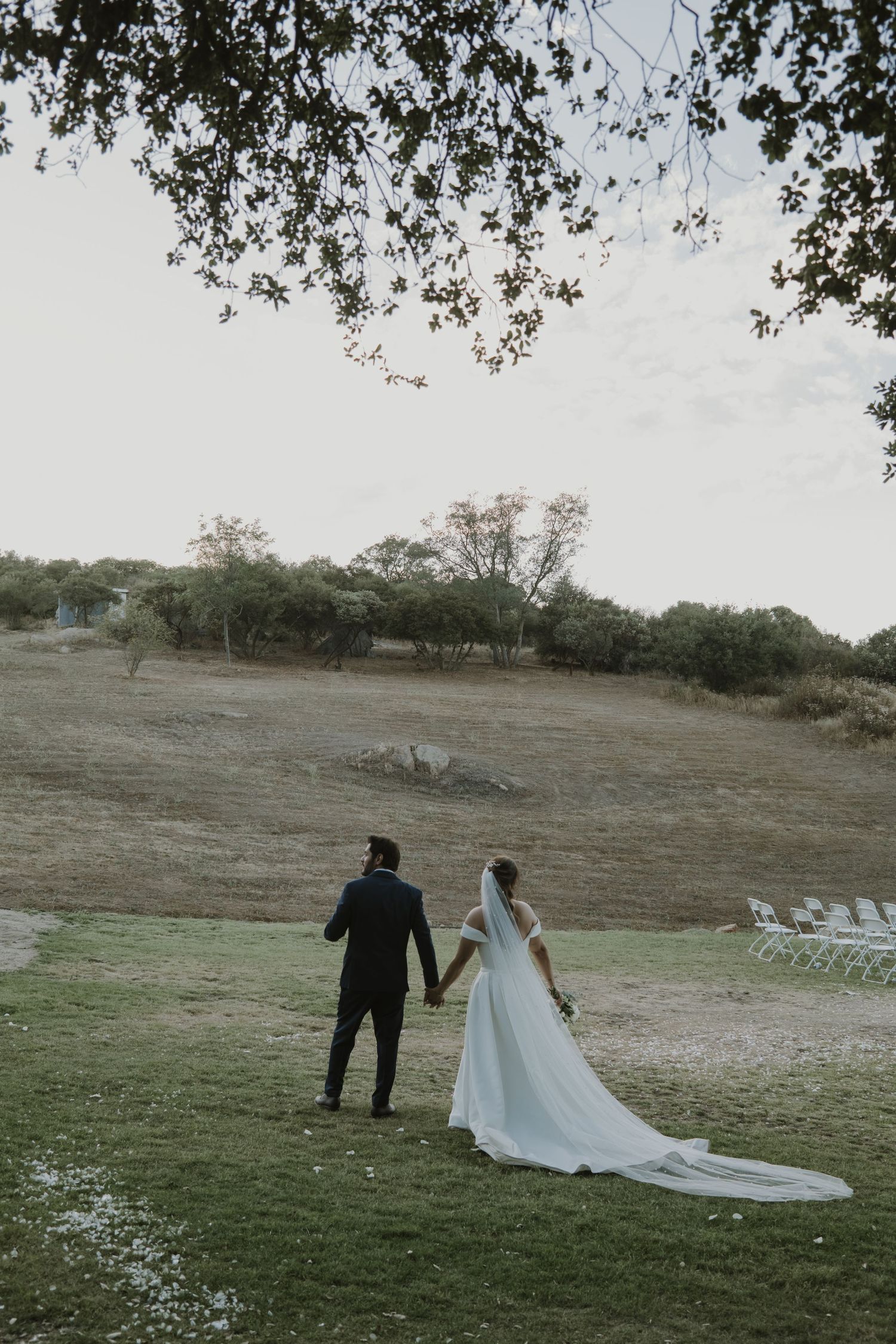 Bride and groom walk hand in hand across a green hillside at their outdoor wedding venue.