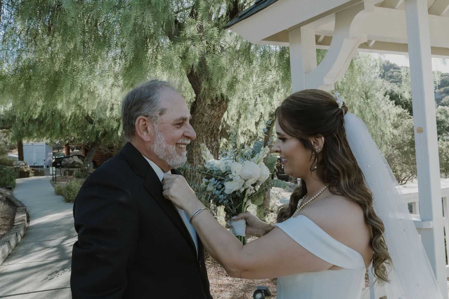 Bride adjusting boutonniere on father's lapel during emotional first look at outdoor wedding ceremony.