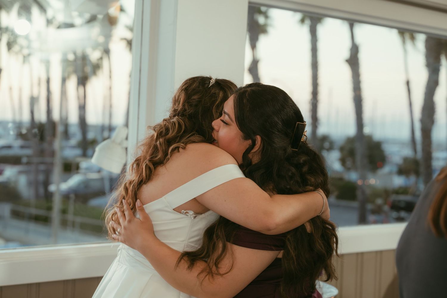 Two women sharing an emotional hug at a wedding with palm trees visible through the window.