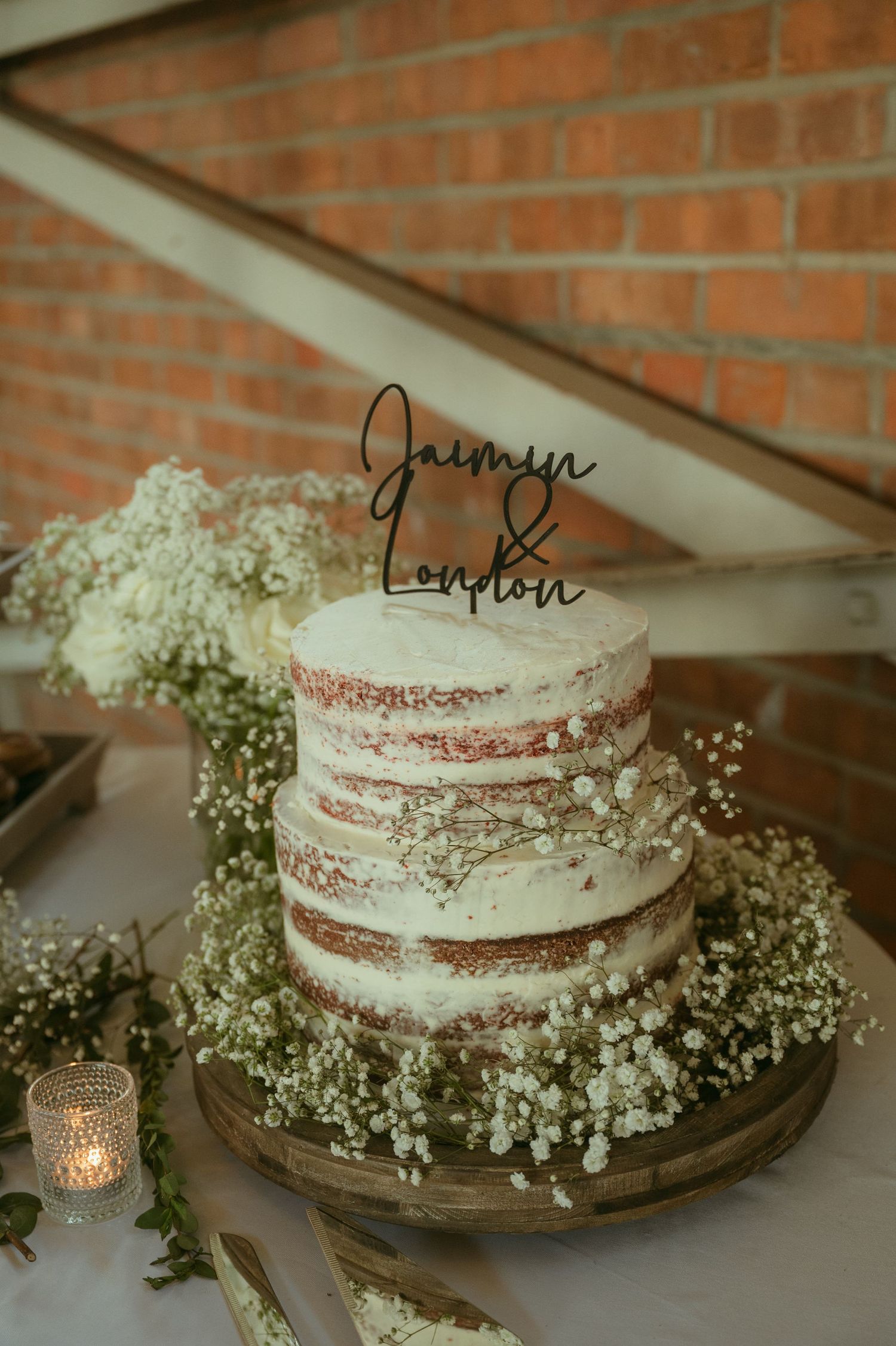Rustic naked wedding cake with baby's breath flowers and custom cake topper reading Jaimyn & London on wooden stand.