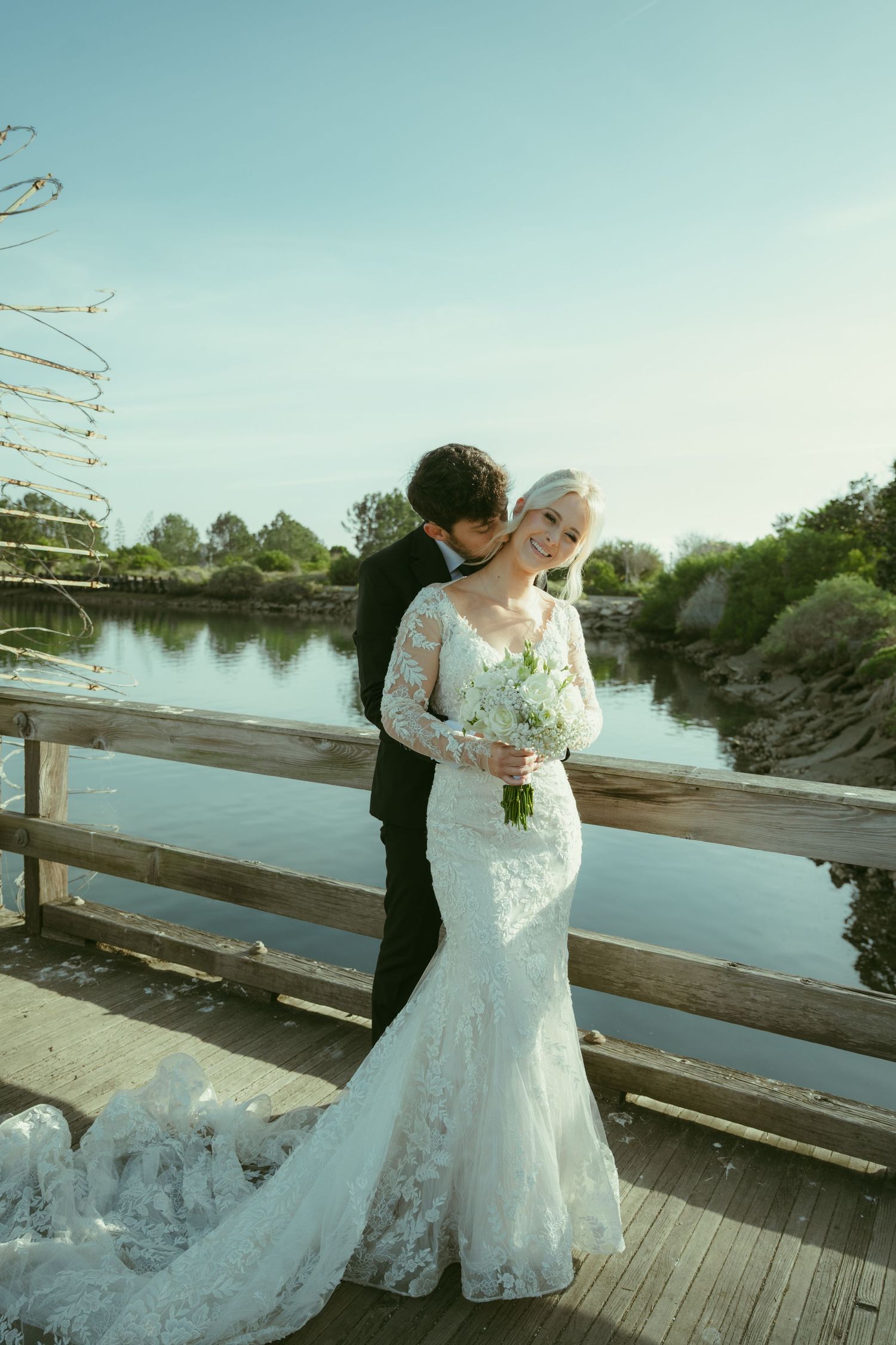 Bride in lace mermaid gown holds white bouquet as groom kisses her cheek on wooden bridge by river.