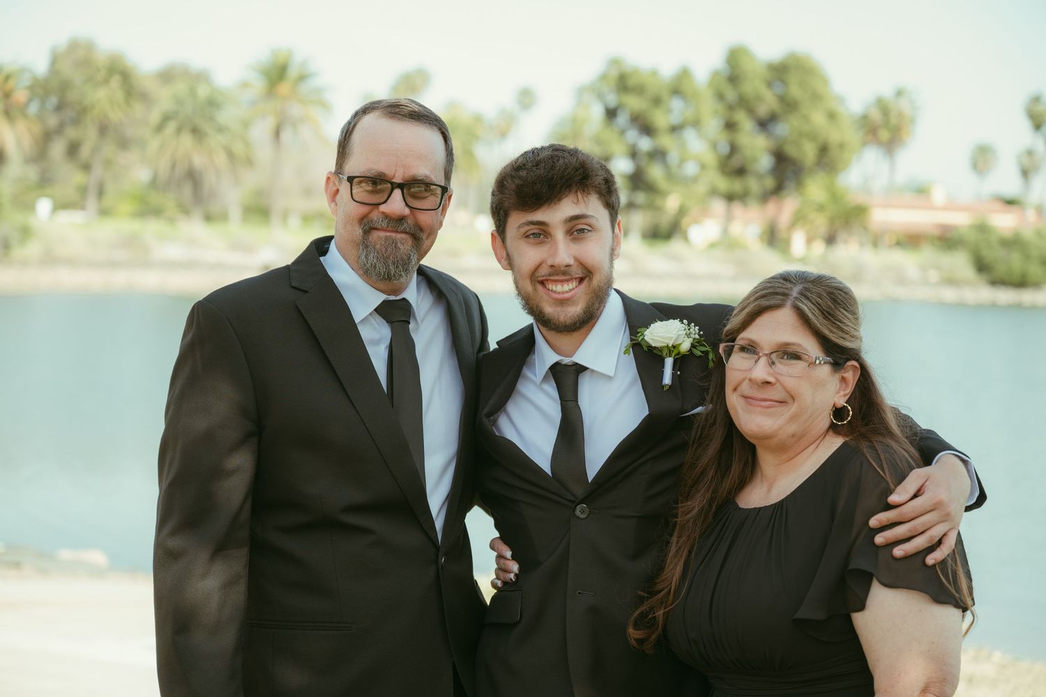 Family of three posing outdoors near a lake, dressed in formal attire at a wedding celebration.
