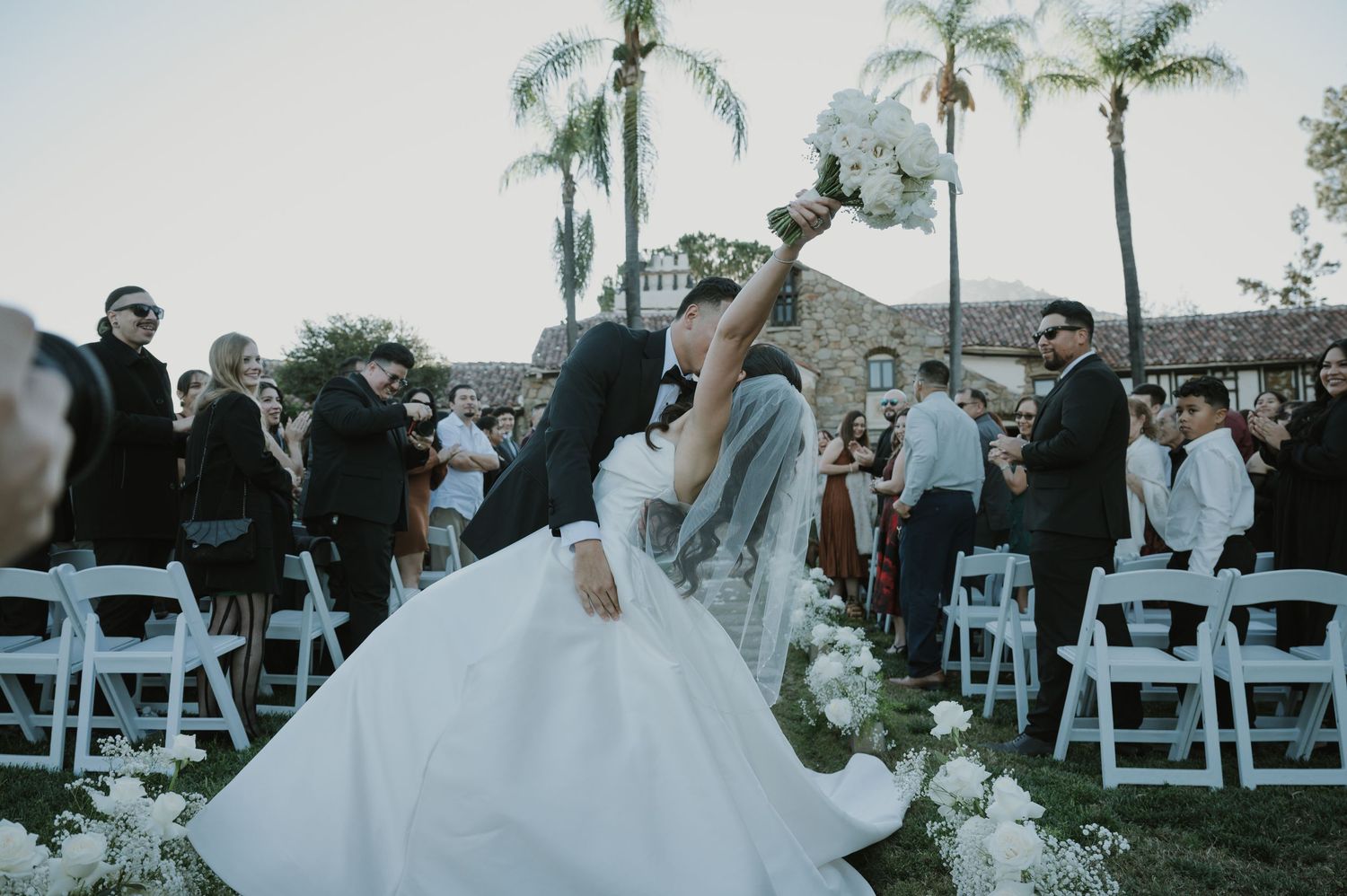 A joyful celebration at an outdoor wedding ceremony with palm trees and white chairs.