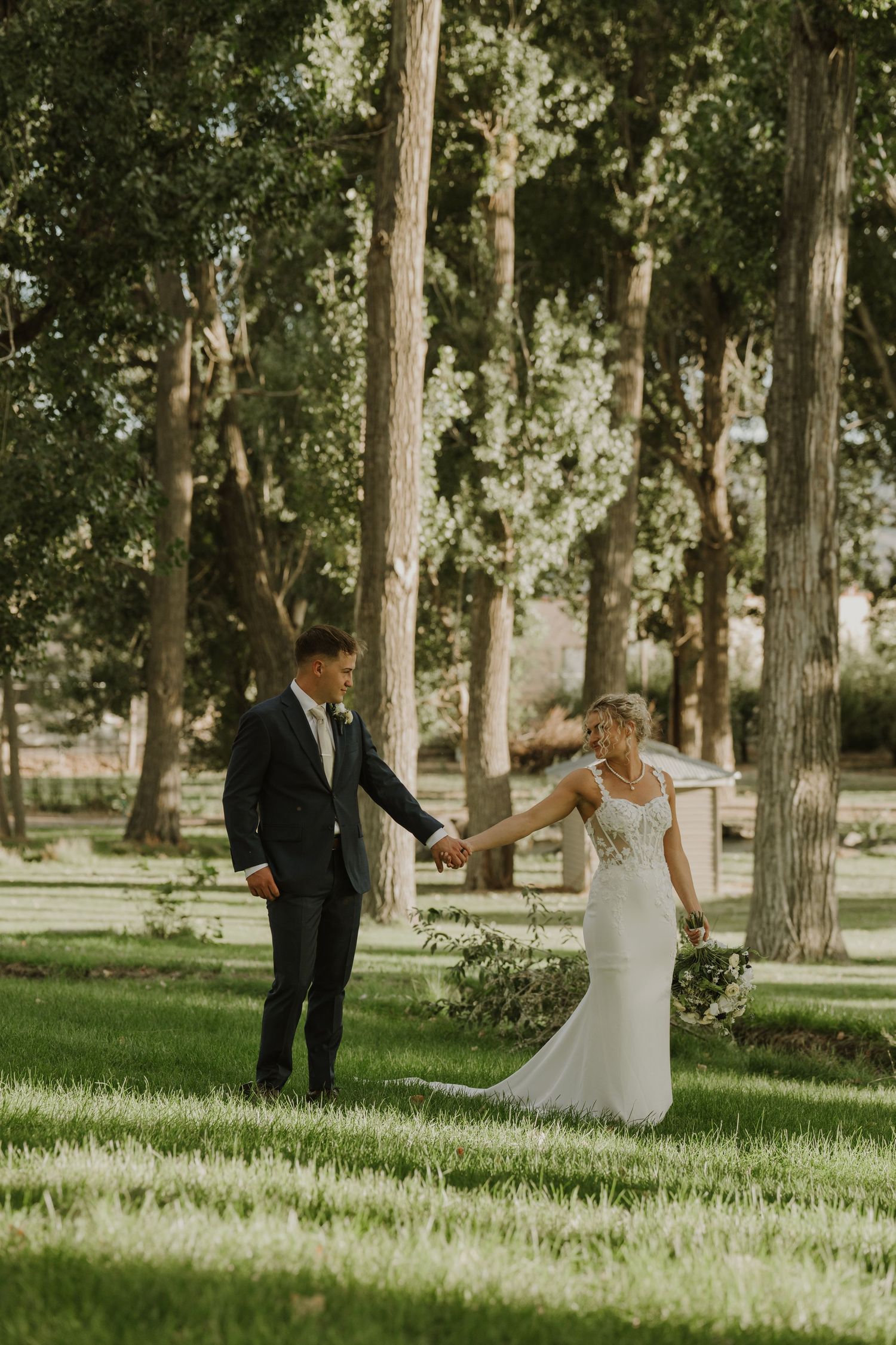 Newlyweds share a romantic moment while walking through a sunlit tree-lined path.