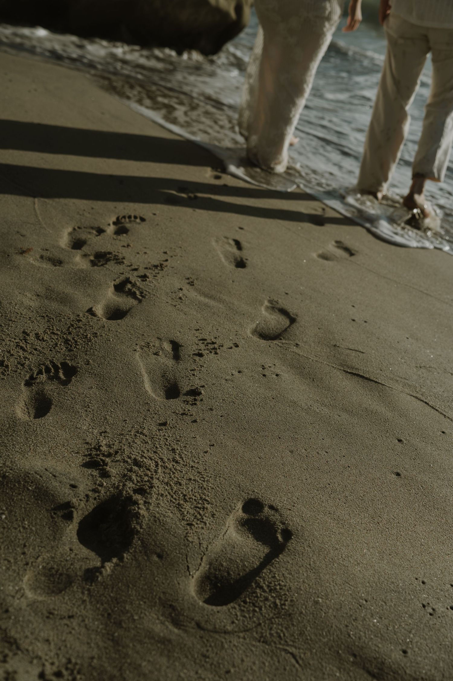 Footprints in dark sand with ocean waves and feet visible in the background.