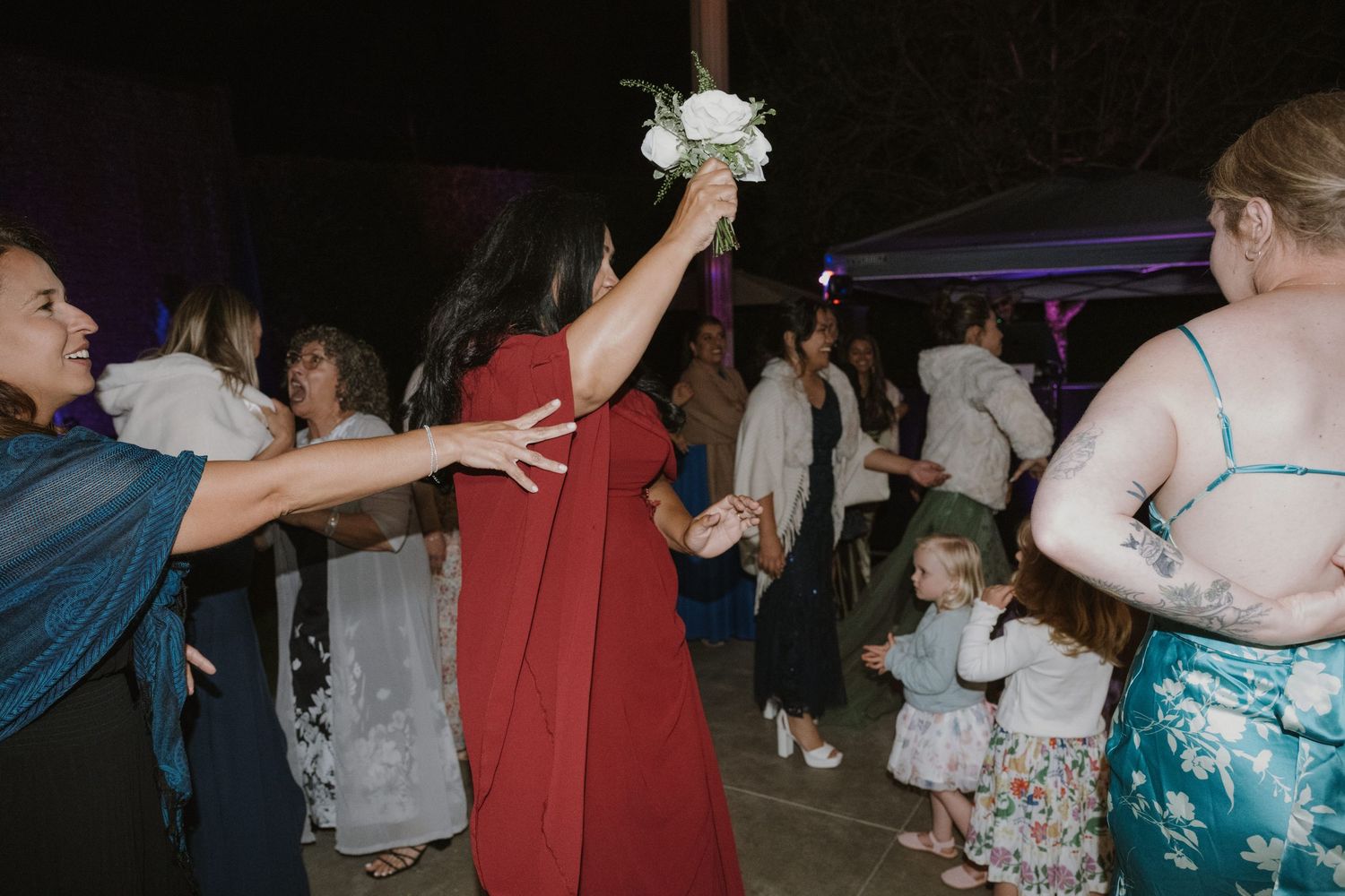 Wedding guest in red dress tossing a white bouquet during the bouquet toss at an outdoor reception.