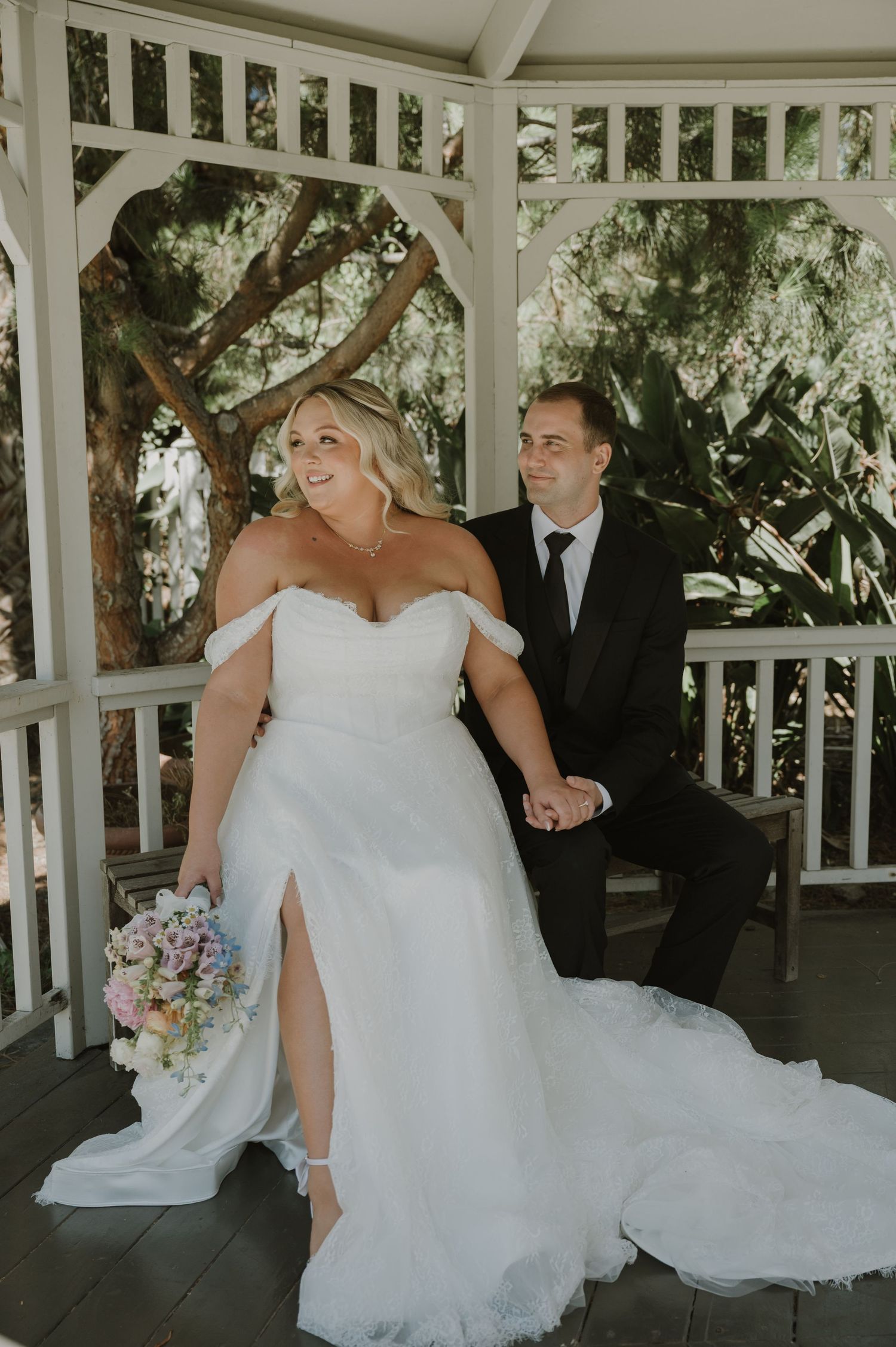 Bride in off-shoulder white gown holds bouquet beside groom in black suit inside white gazebo.
