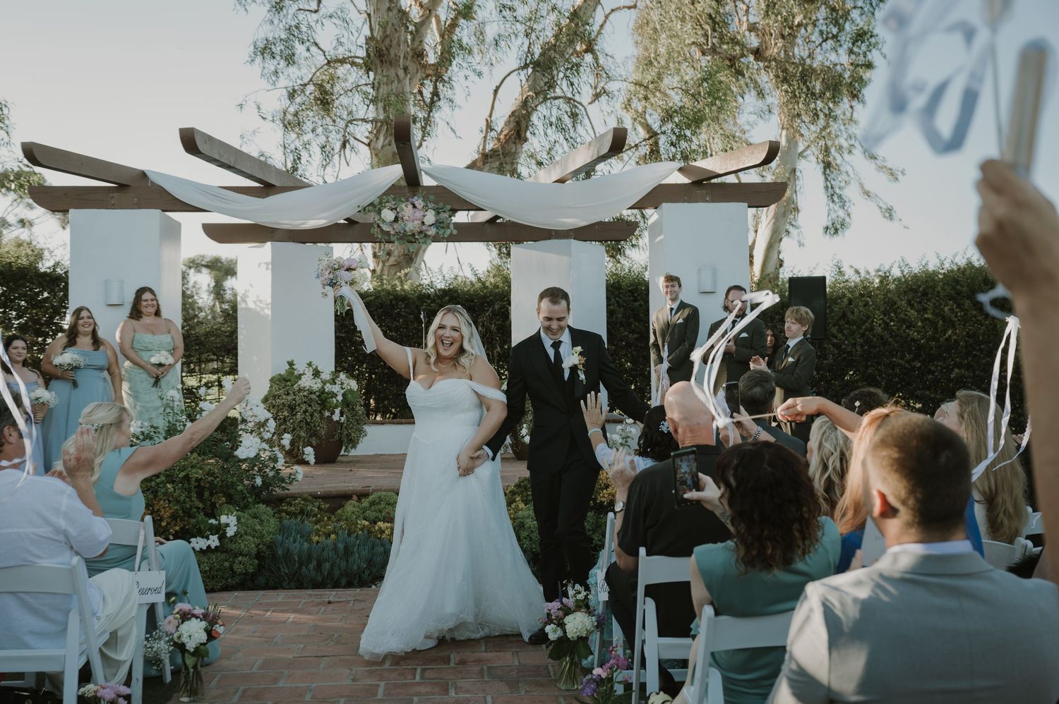 Newlyweds celebrate recessional as guests wave ribbons at elegant outdoor garden wedding ceremony.