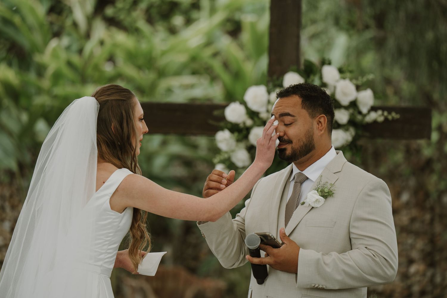 Bride gently wipes groom's tear during outdoor wedding ceremony near floral cross.