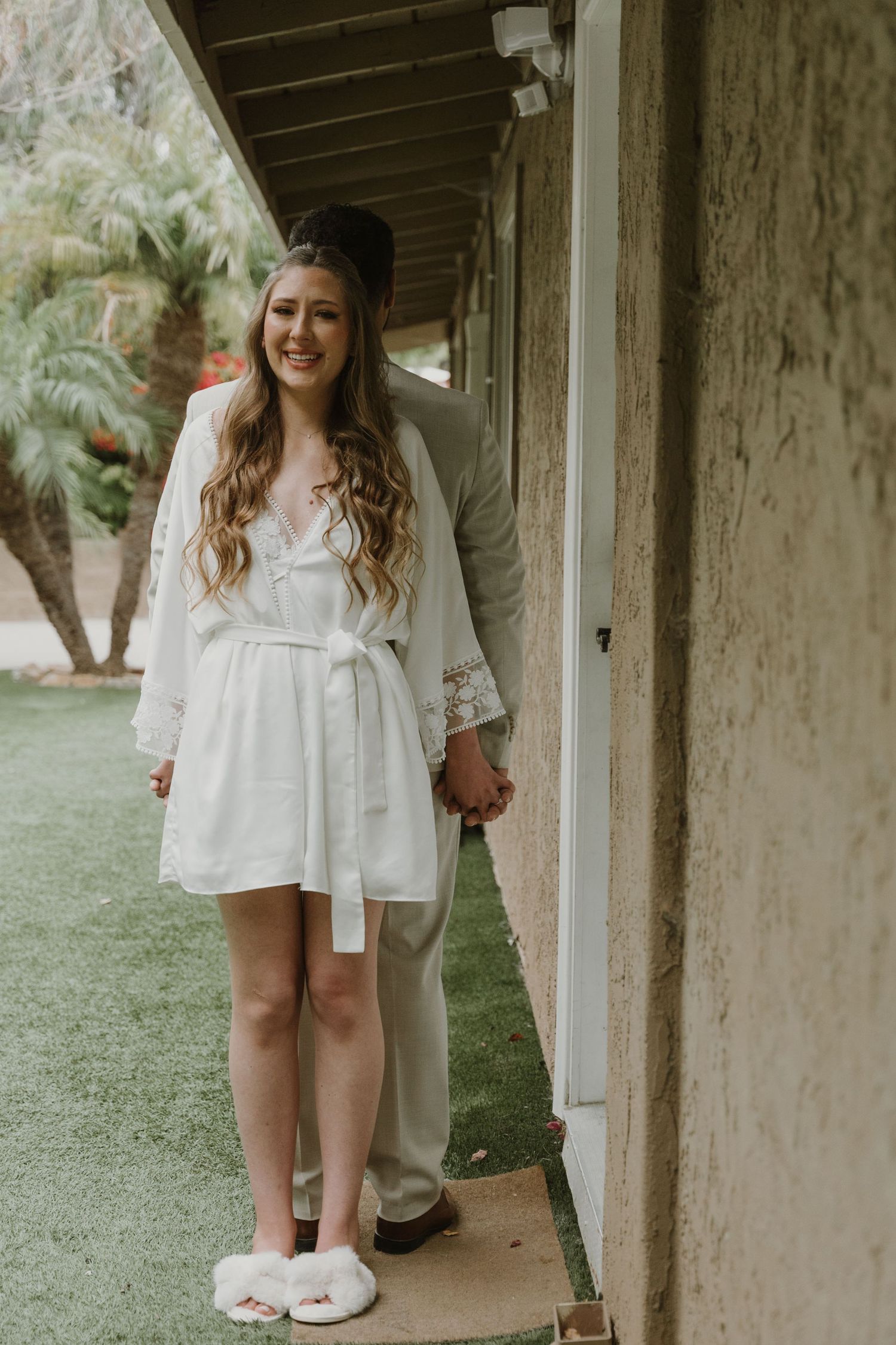 Smiling bride in white lace robe and fluffy slippers holds hands with groom in beige suit outside a venue.