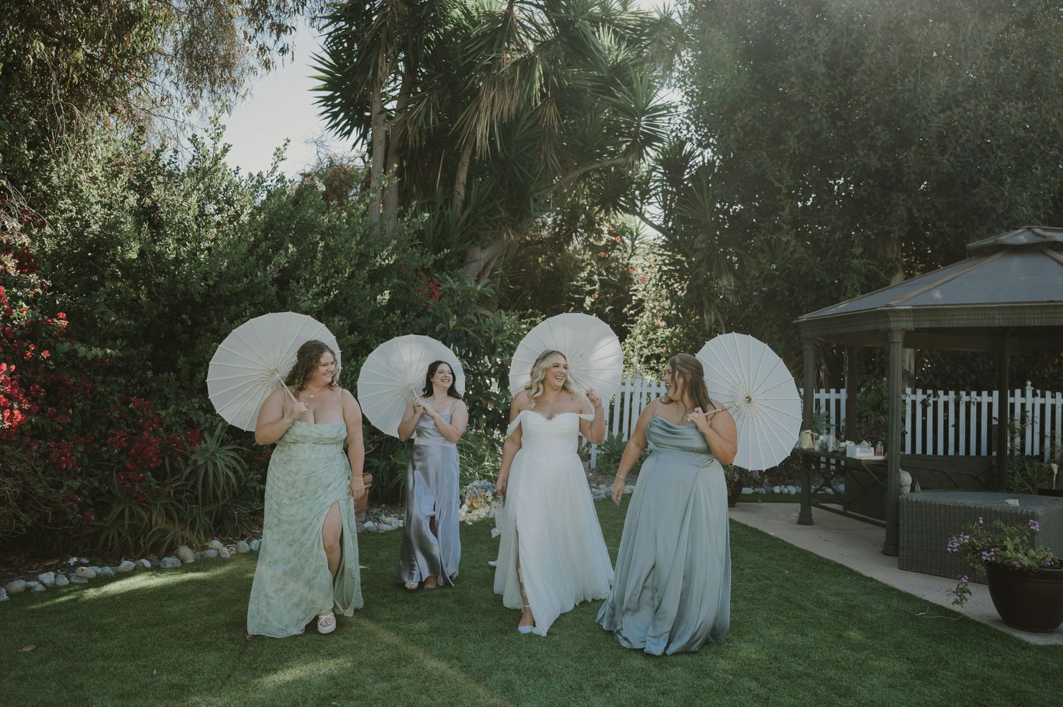 Bride and bridesmaids walking in garden holding white parasols, wearing mismatched pastel gowns outdoors.
